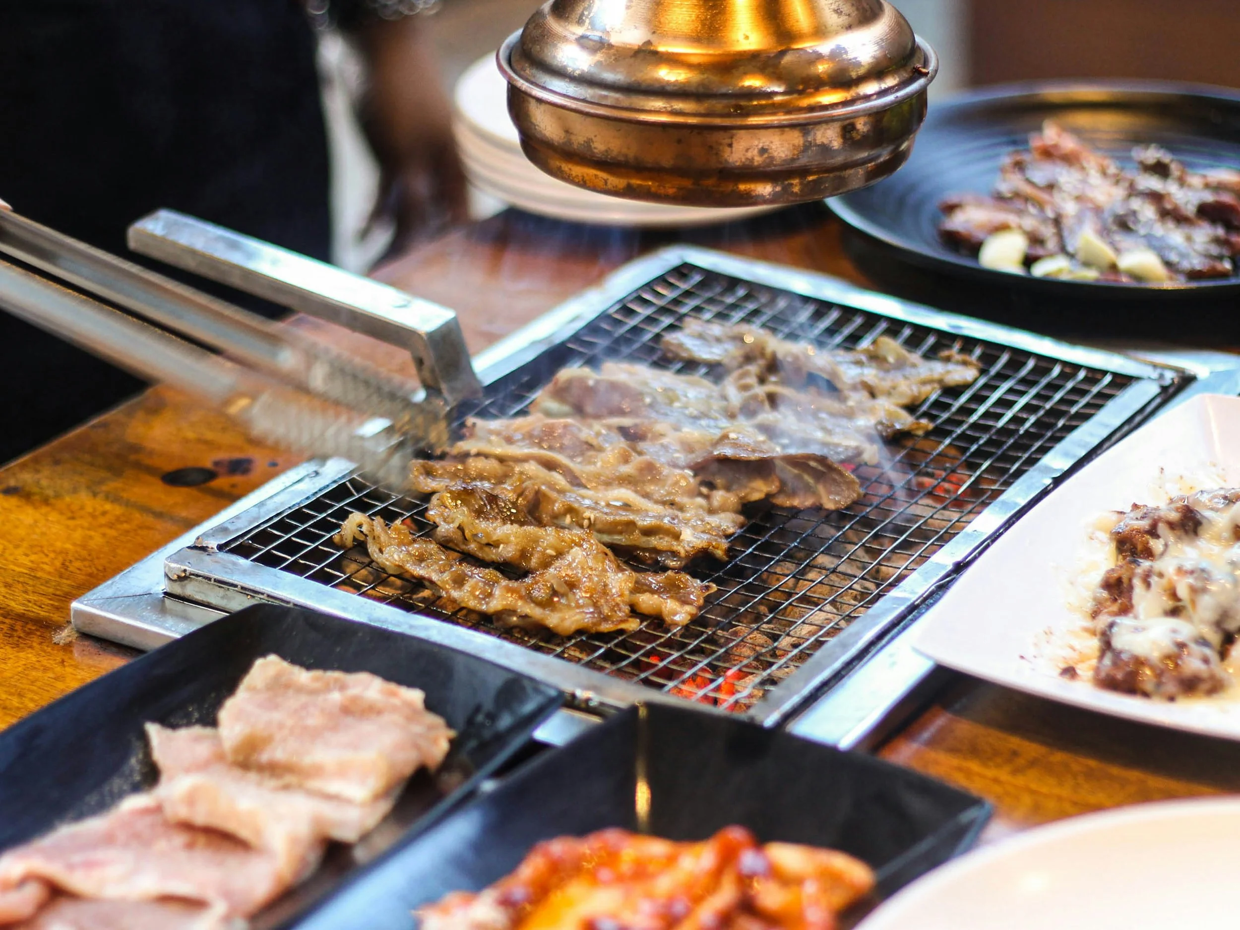 Cooking meat on a barbecue grill with tongs, with plates of raw and cooked meat on a wooden table.