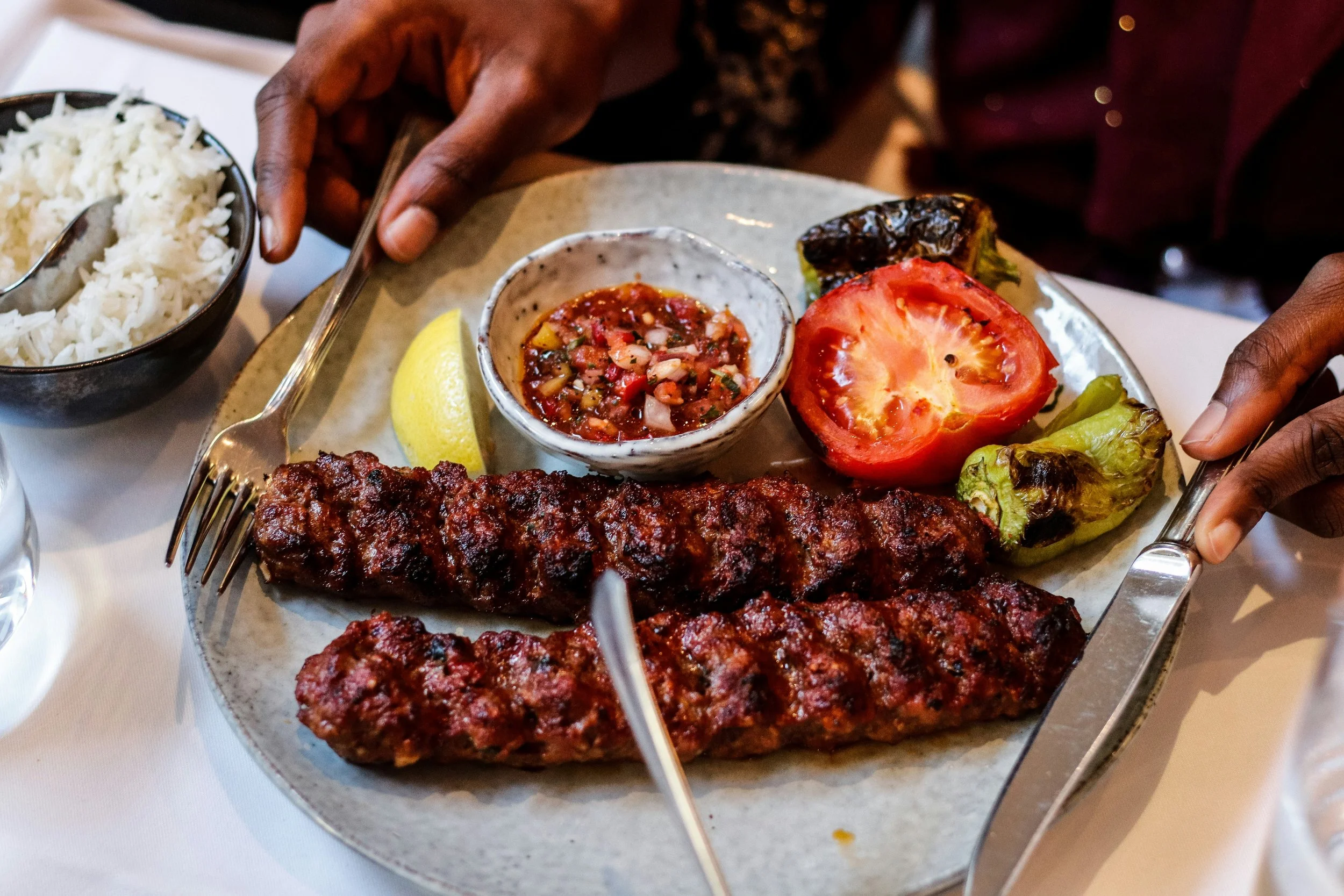 Plate of grilled meat skewers, fresh tomato slice, grilled peppers, lemon wedge, side of white rice, and a small bowl of tomato-based sauce with chopped vegetables.