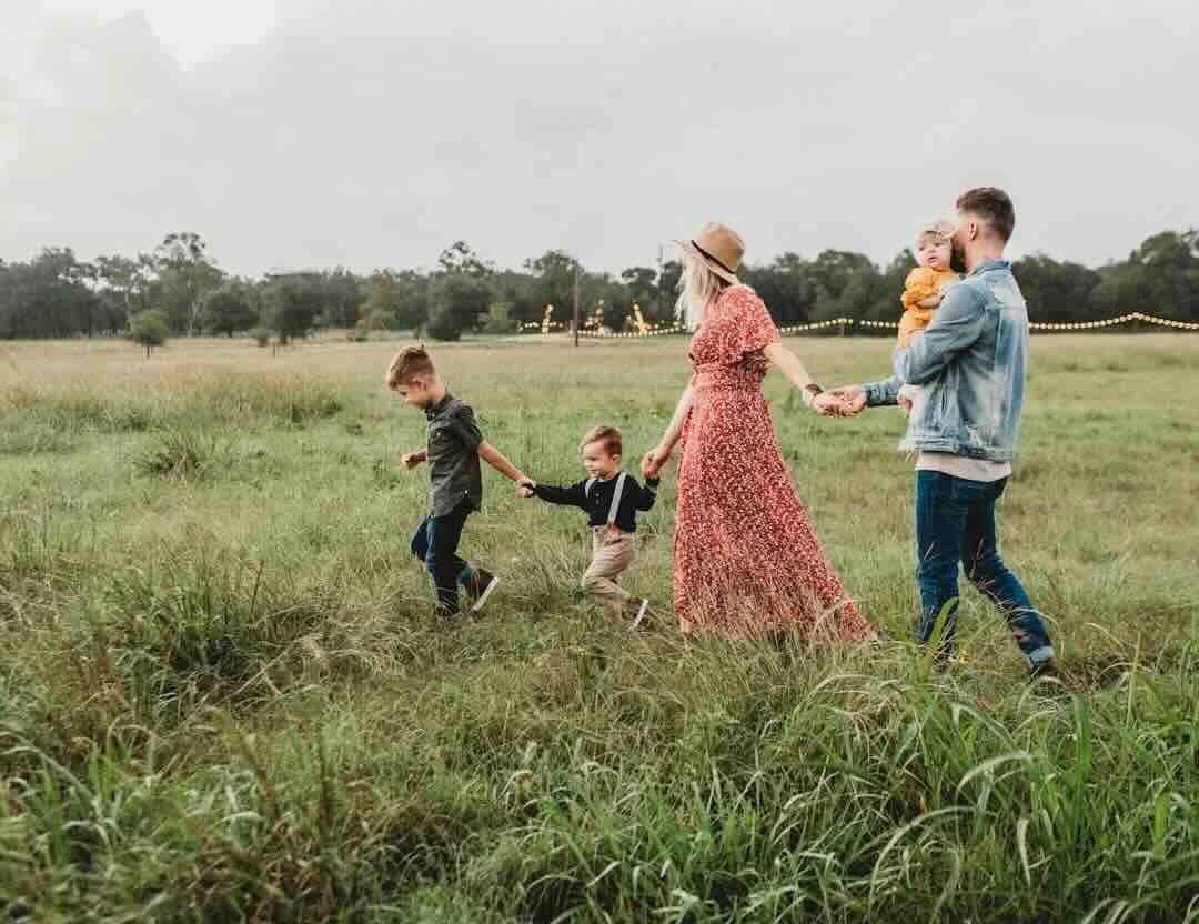 Family walking in a grassy field with kids holding hands, overcast sky, some trees and string lights in the background.