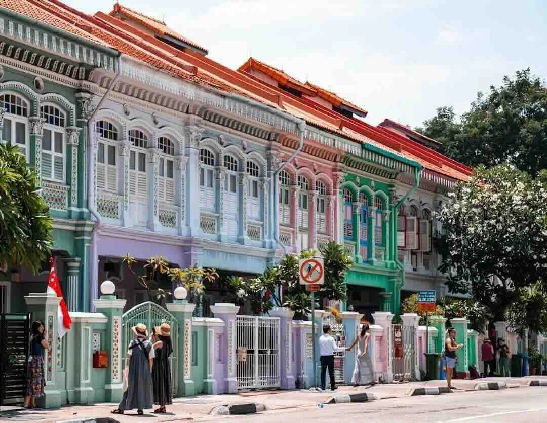 Colorful row of Peranakan shophouses with decorative facades in pastel shades, people walking on the sidewalk, trees lining the street, and a sign indicating no parking.
