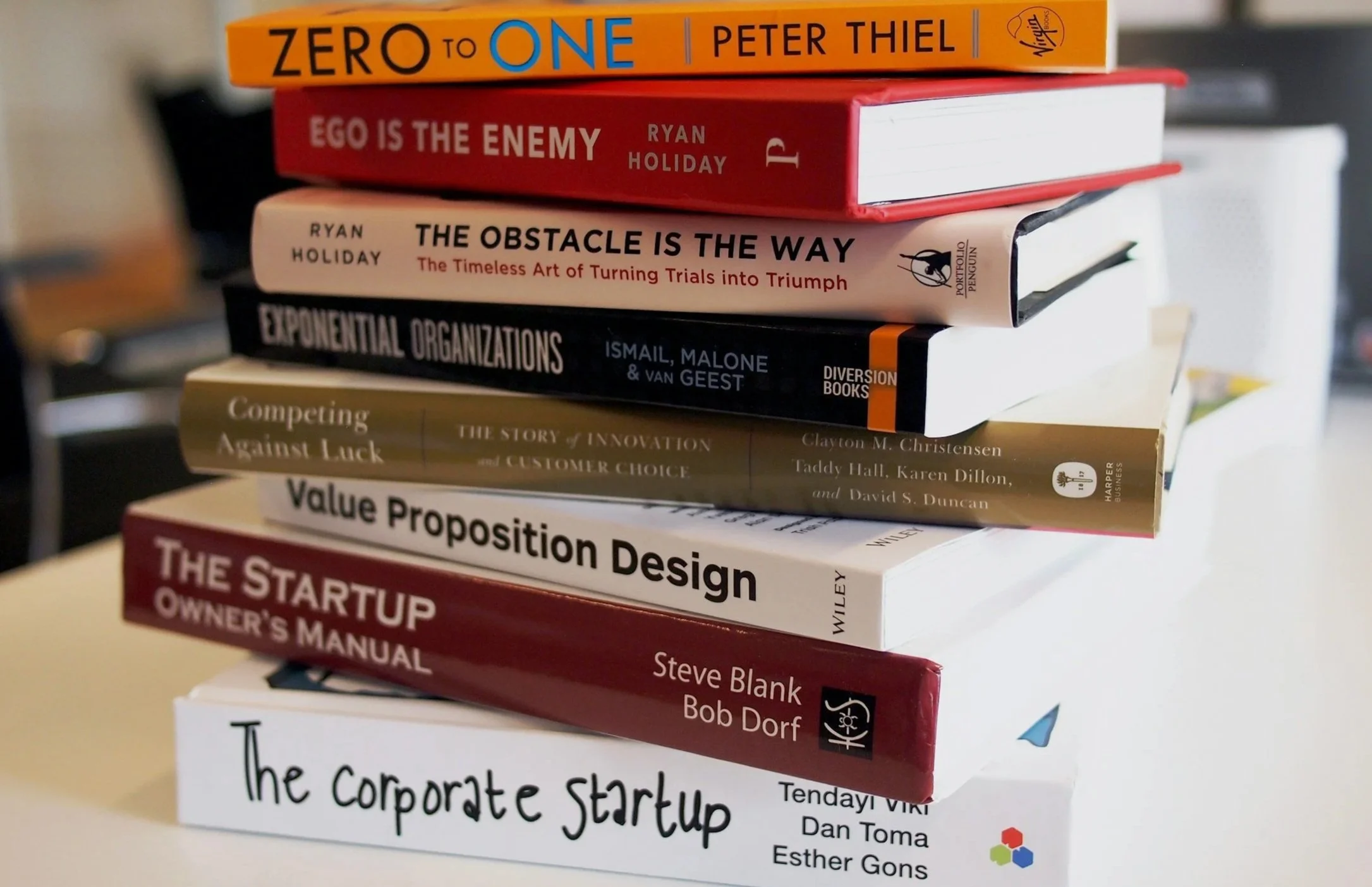 Stack of several colorful books with titles including 'Zero to One' by Peter Thiel, 'Ego is the Enemy' by Ryan Holiday, 'The Obstacle is the Way' by Ryan Holiday, 'Exponential Organizations' by Ismail, Malone & Van Geest, 'Competing Against Luck,' 'Value Proposition Design,' 'The Startup Owner's Manual,' and 'The Corporate Startup' by Tendayi Viki, Dan Toma, and Esther Gons.
