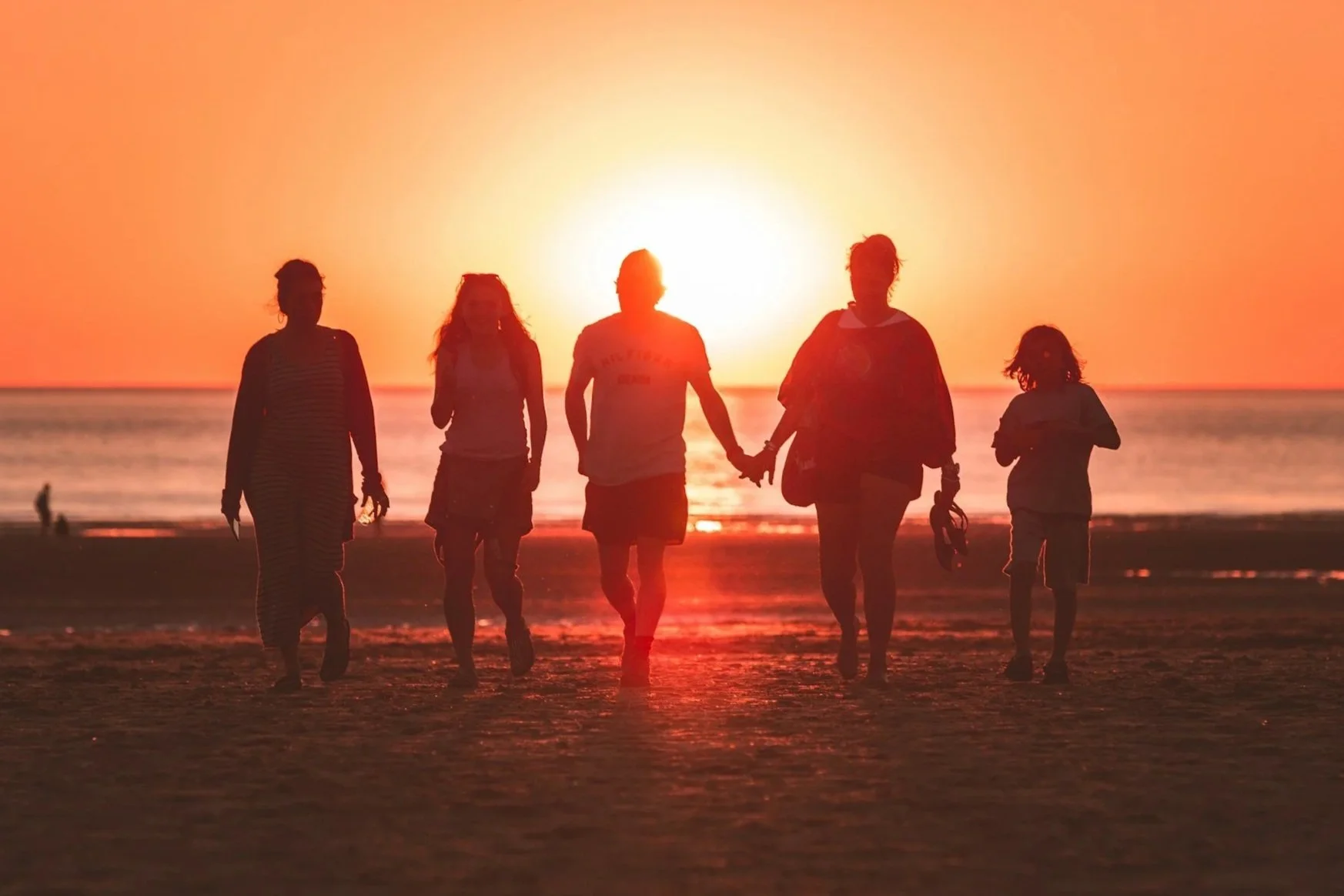 Silhouettes of a family walking along the beach at sunset, holding hands, with the ocean in the background and the sun low on the horizon.