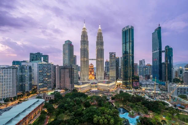 City skyline with tall skyscrapers, including the Petronas Towers, and a park with trees and a pool in the foreground during dusk.