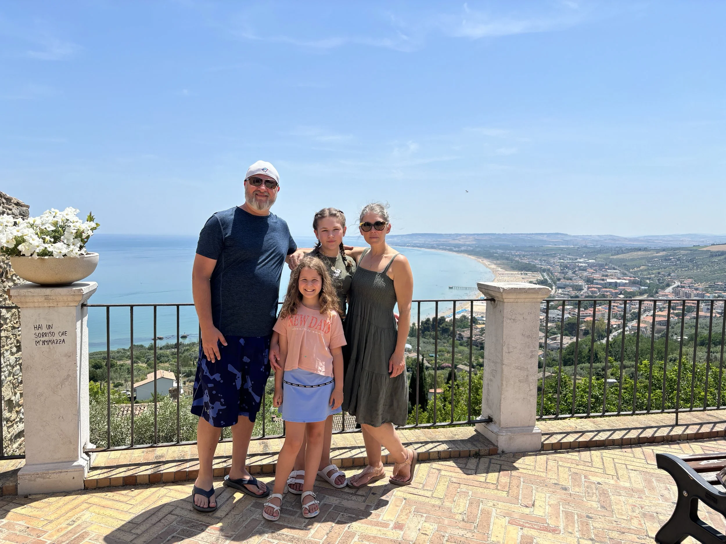 Family of four standing on a viewing platform overlooking a coastal city with a beach, water, and hills in the background on a sunny day.