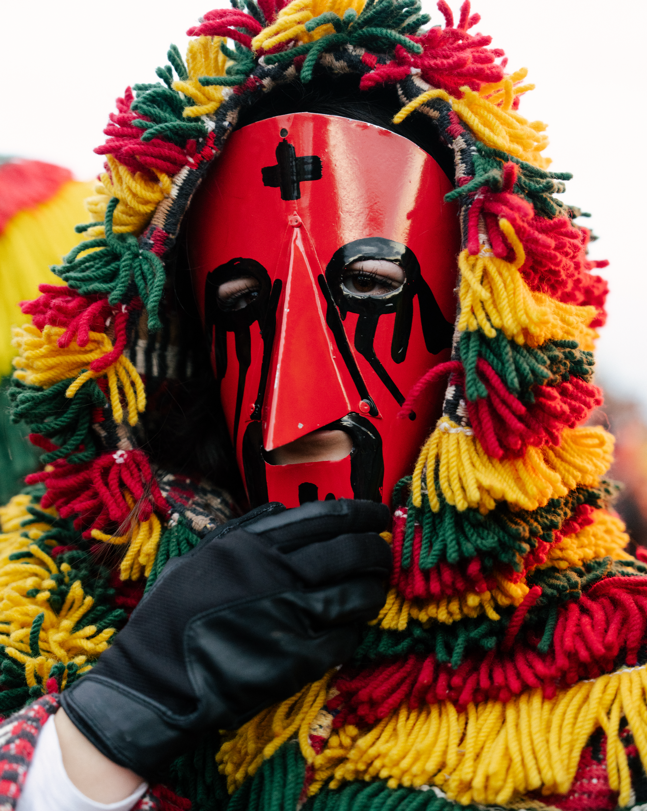 Person wearing a red mask with black and white designs and a colorful scarf made of thick yarn in red, yellow, green, and black, with a pattern of intersecting lines. The person is holding the mask with their gloved hand.
