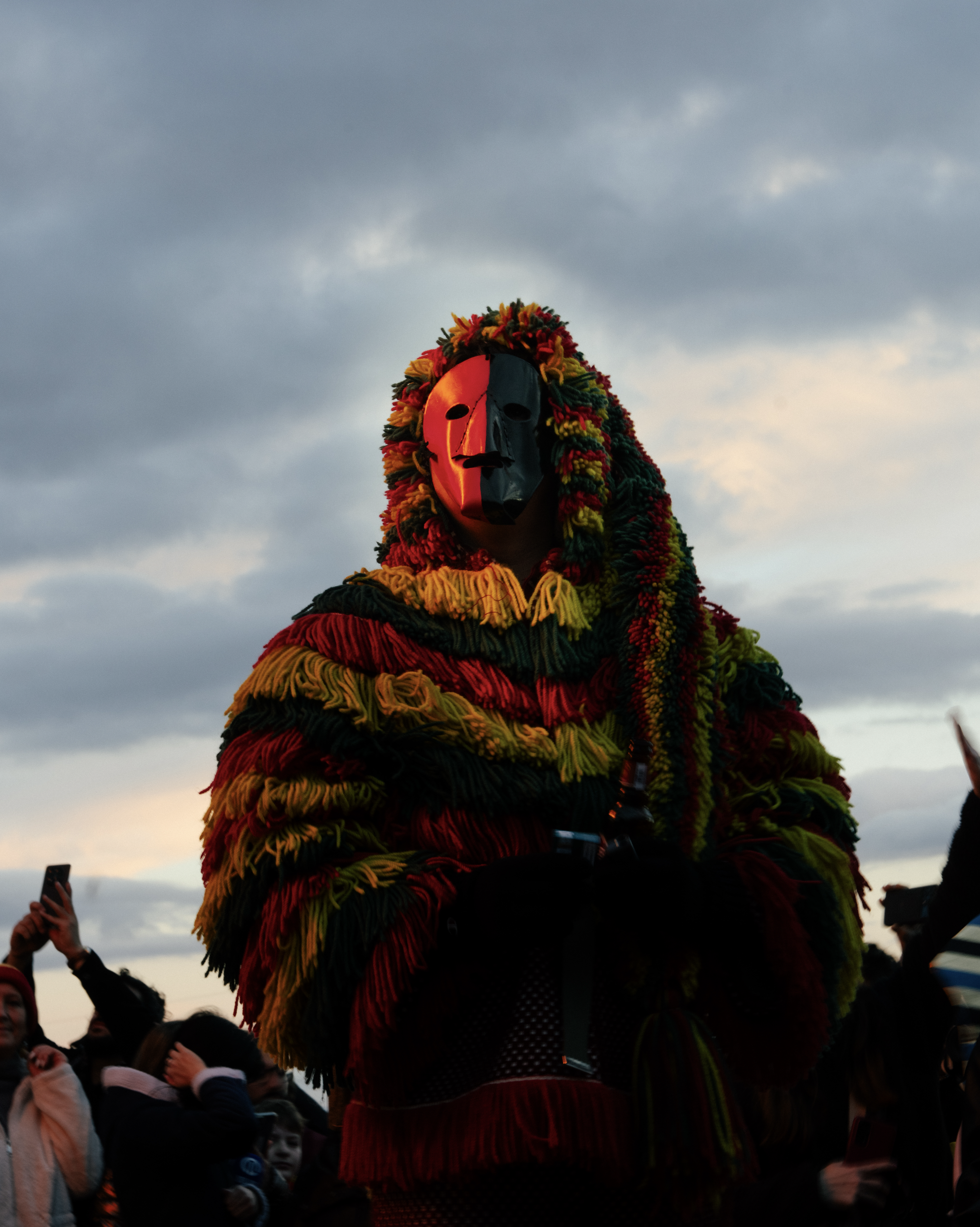 A person wearing a black and red mask with a multicolored yarn headdress during a parade or celebration, surrounded by a crowd of onlookers under an overcast sky.