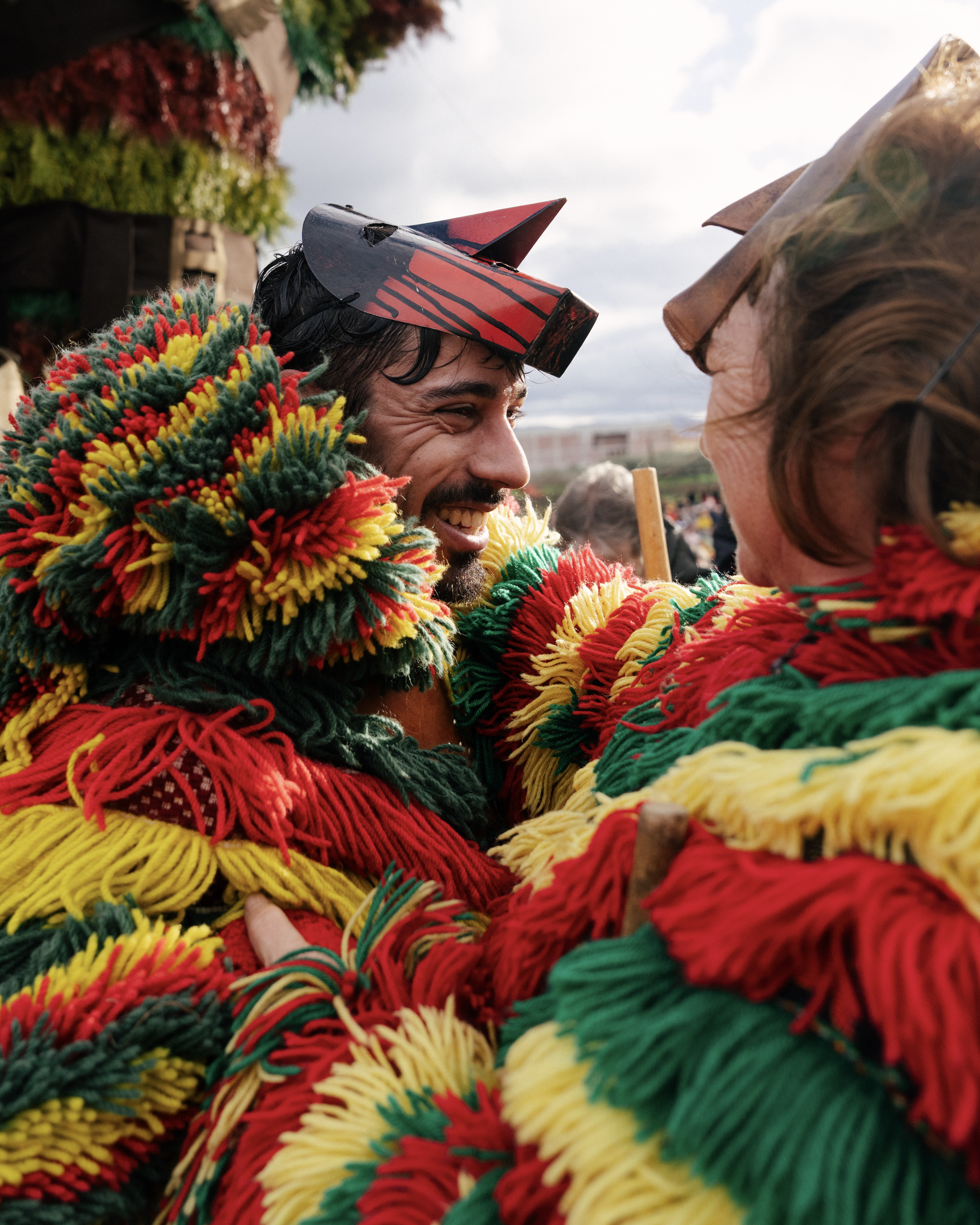 Two people dressed in colorful, thick, yarn-like costumes, smiling at each other, with one wearing a paper hat.