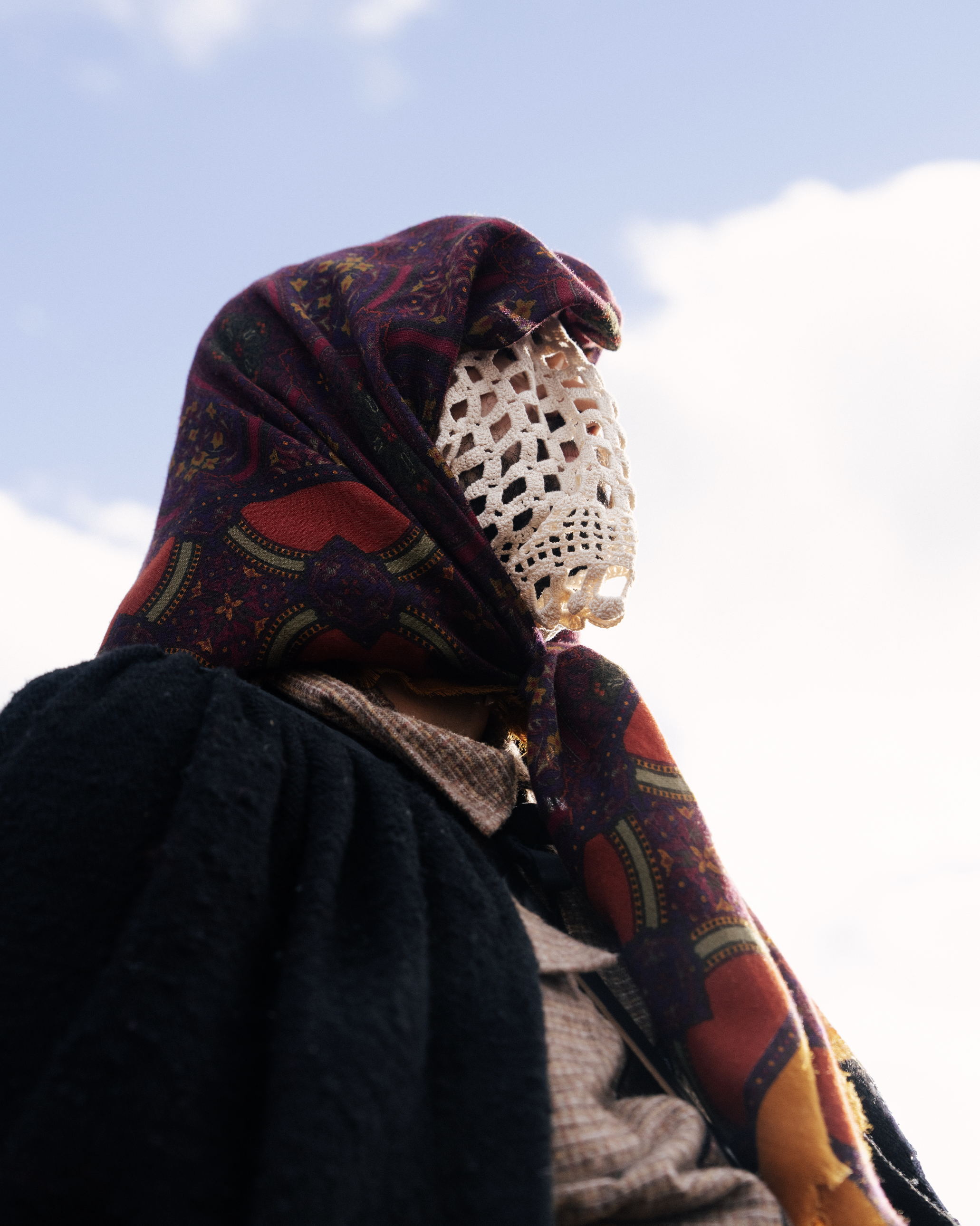 Close-up of a person wrapped in a colorful patterned scarf and crochet face cover, wearing layered clothing, against a bright blue sky with some clouds.