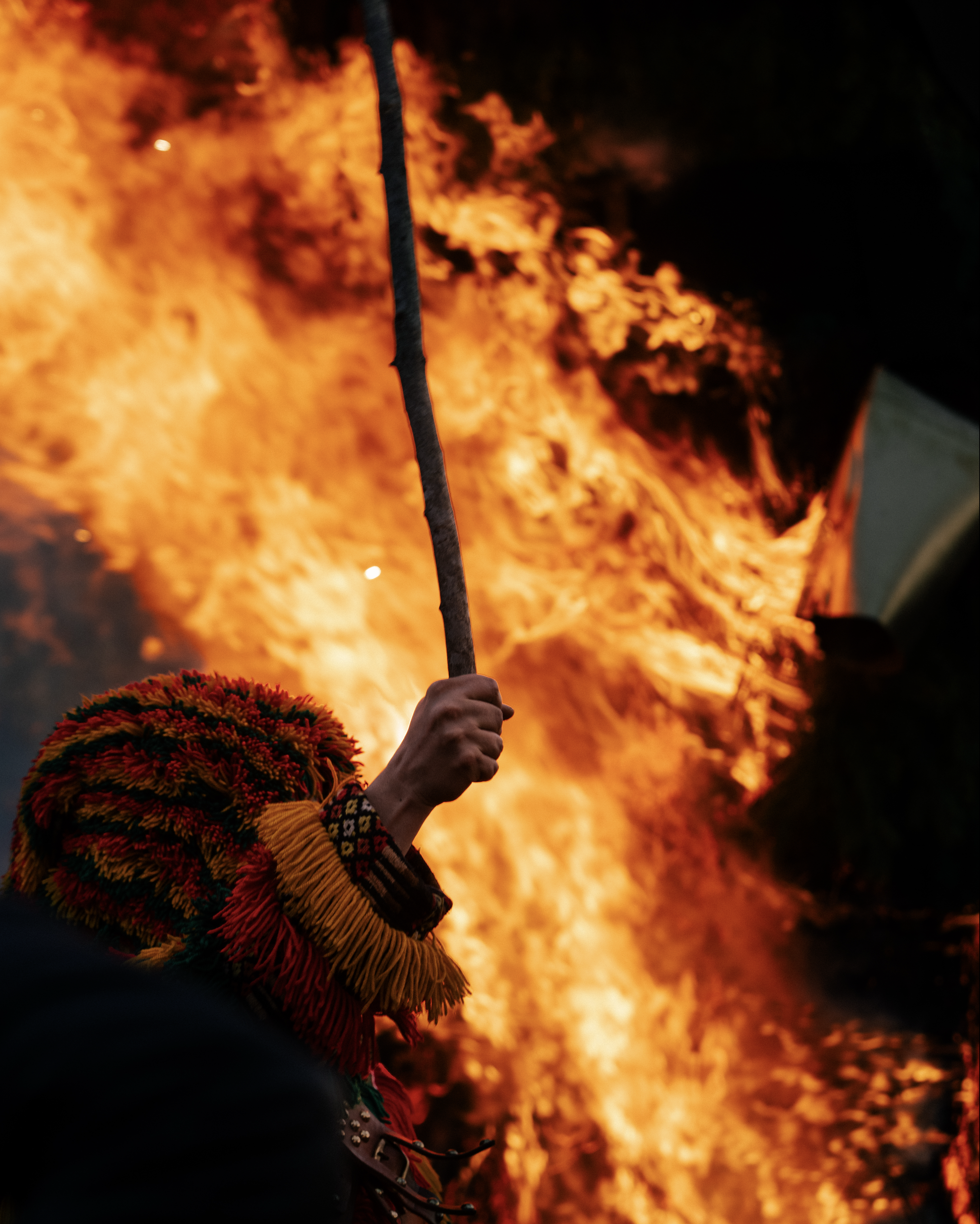 A person holding a lit candle in front of large flames, wearing a colorful, textured costume and floral headwear.