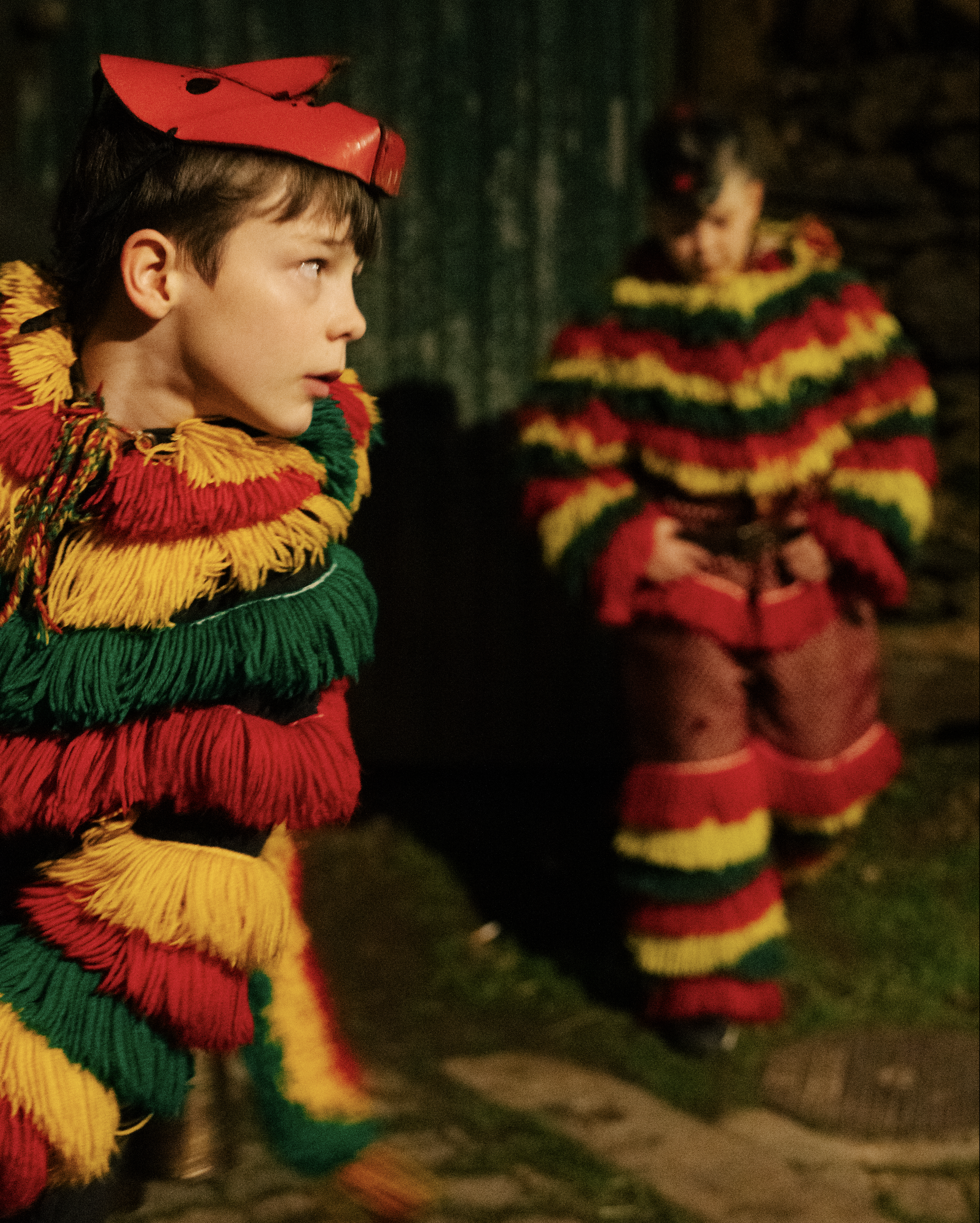 Two children dressed in bright, colorful, striped costumes with fringes, standing outdoors at night.