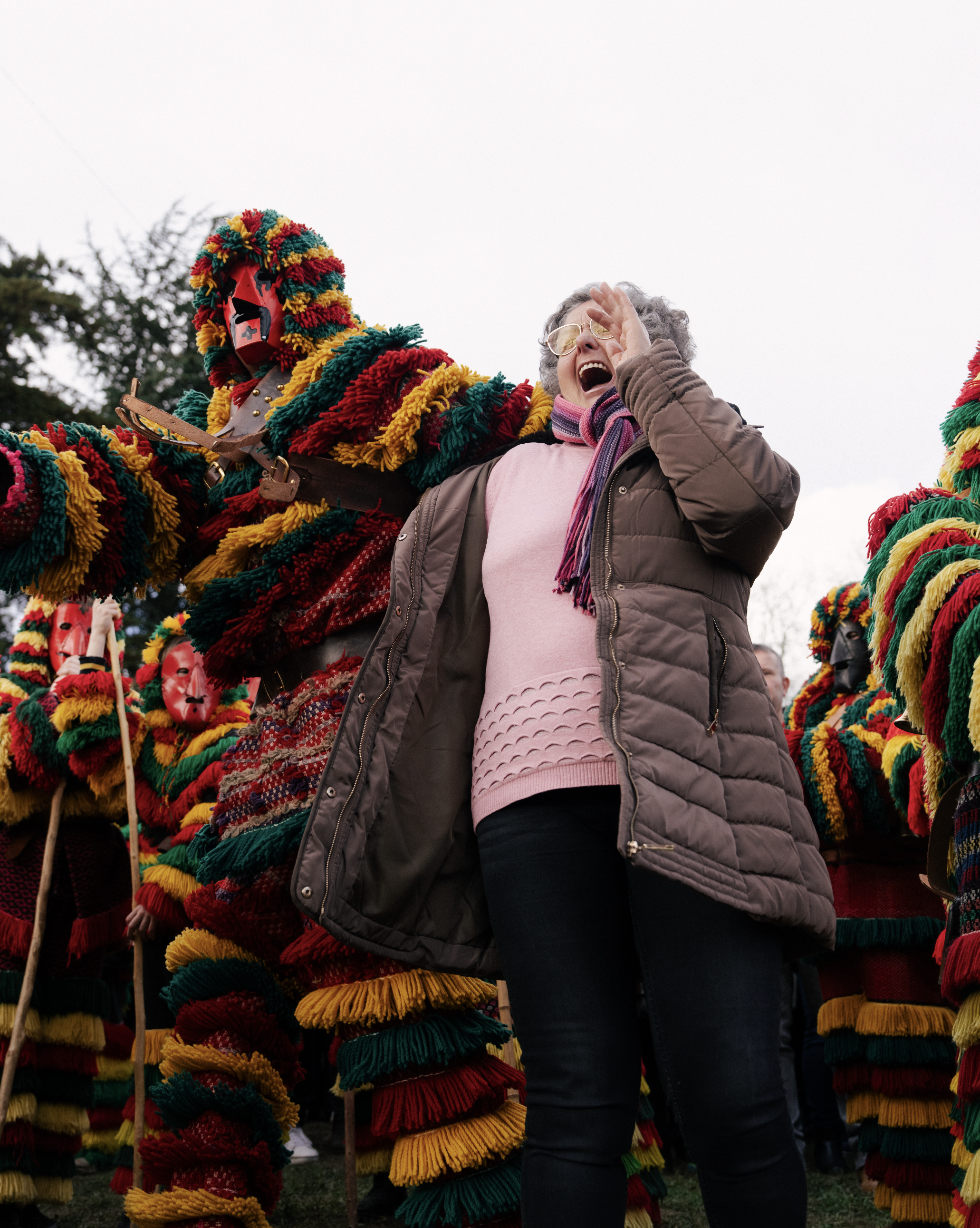 A woman laughing with her hand near her face, standing among costumed performers wearing colorful, striped pom-pom outfits and masks, outdoors during daytime.