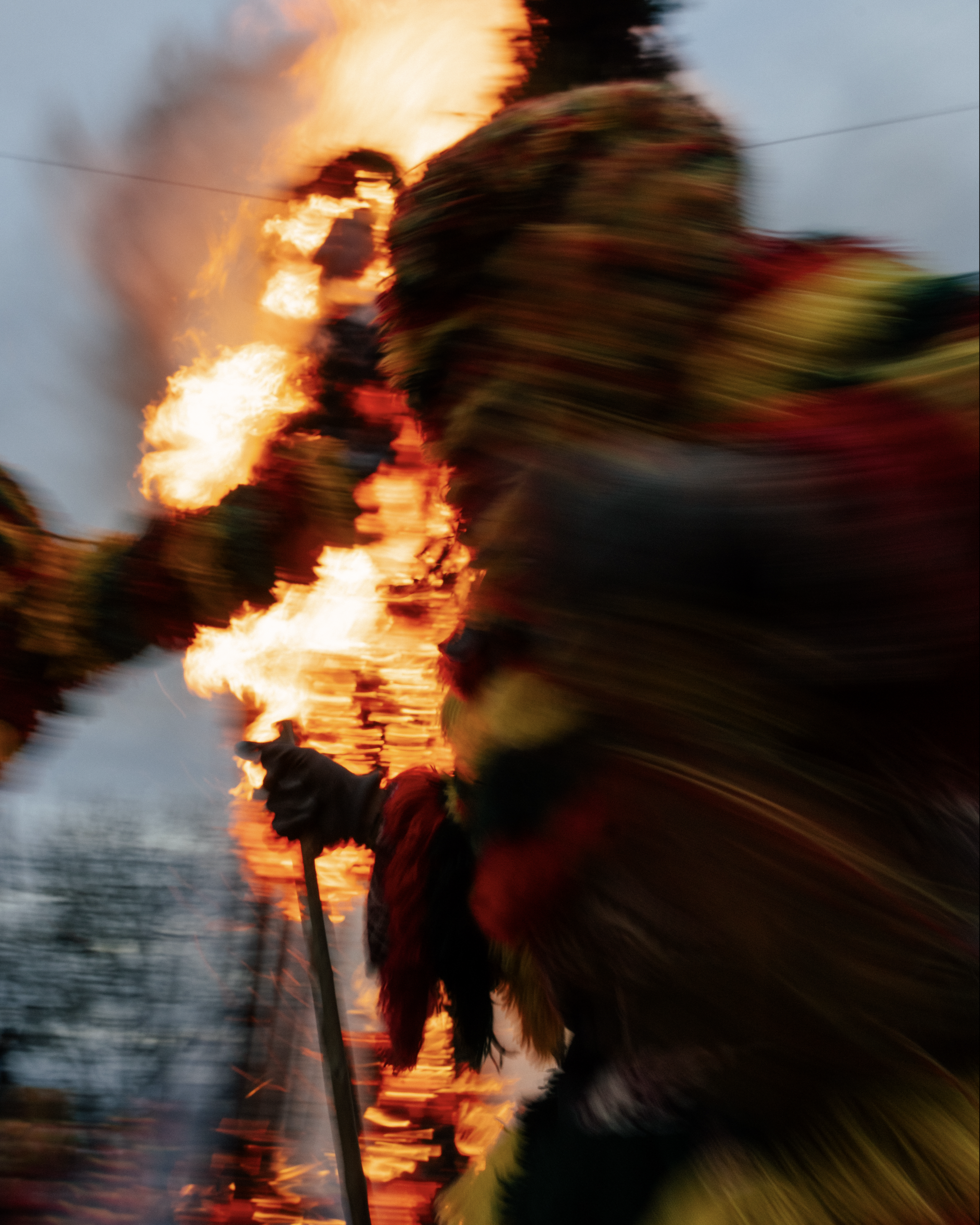A blurry photograph of a person holding a torch on a boat during a sunset, with flames and smoke in the background.