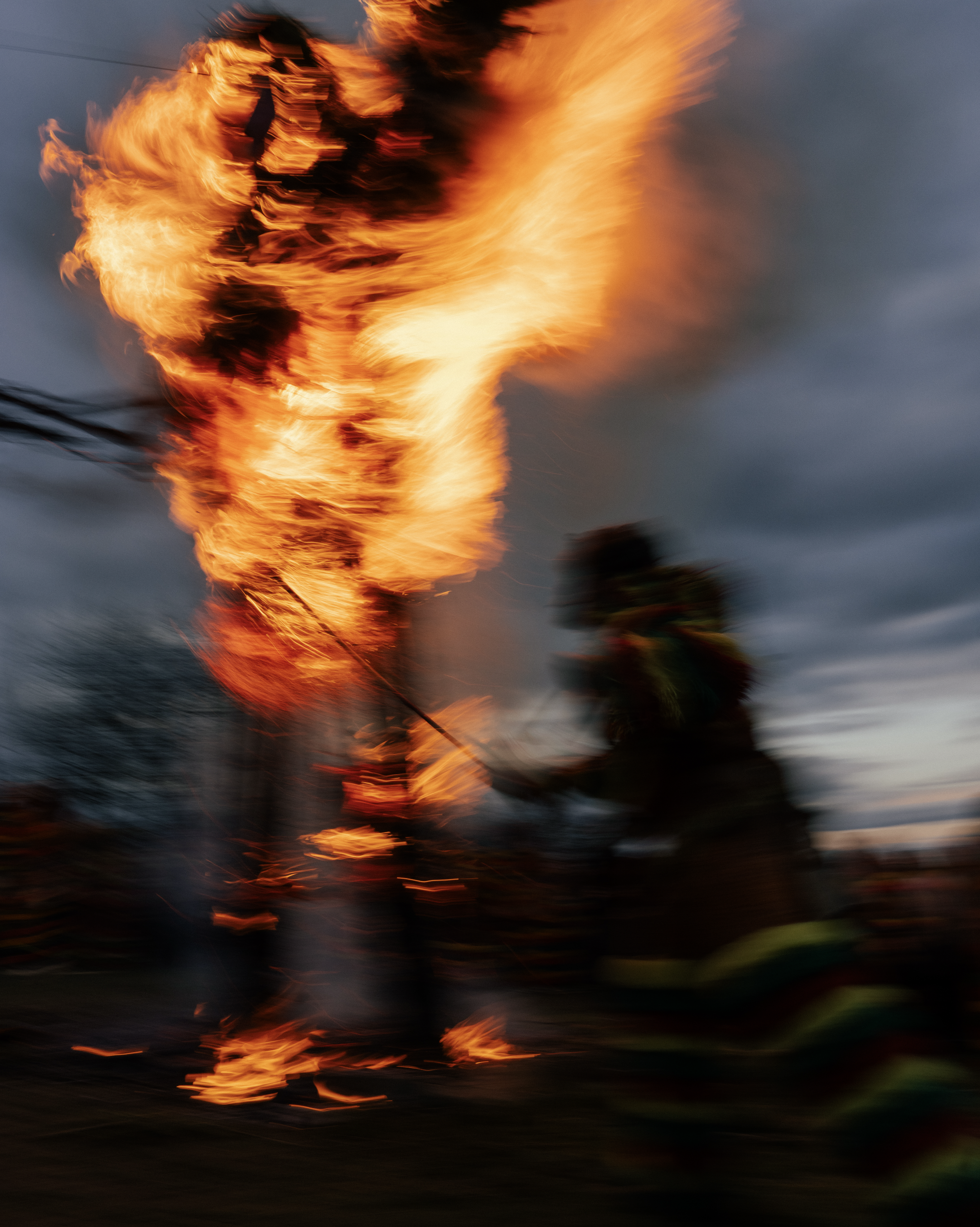 Blurred image of a large fire with intense orange and yellow flames against a dark, cloudy sky background.