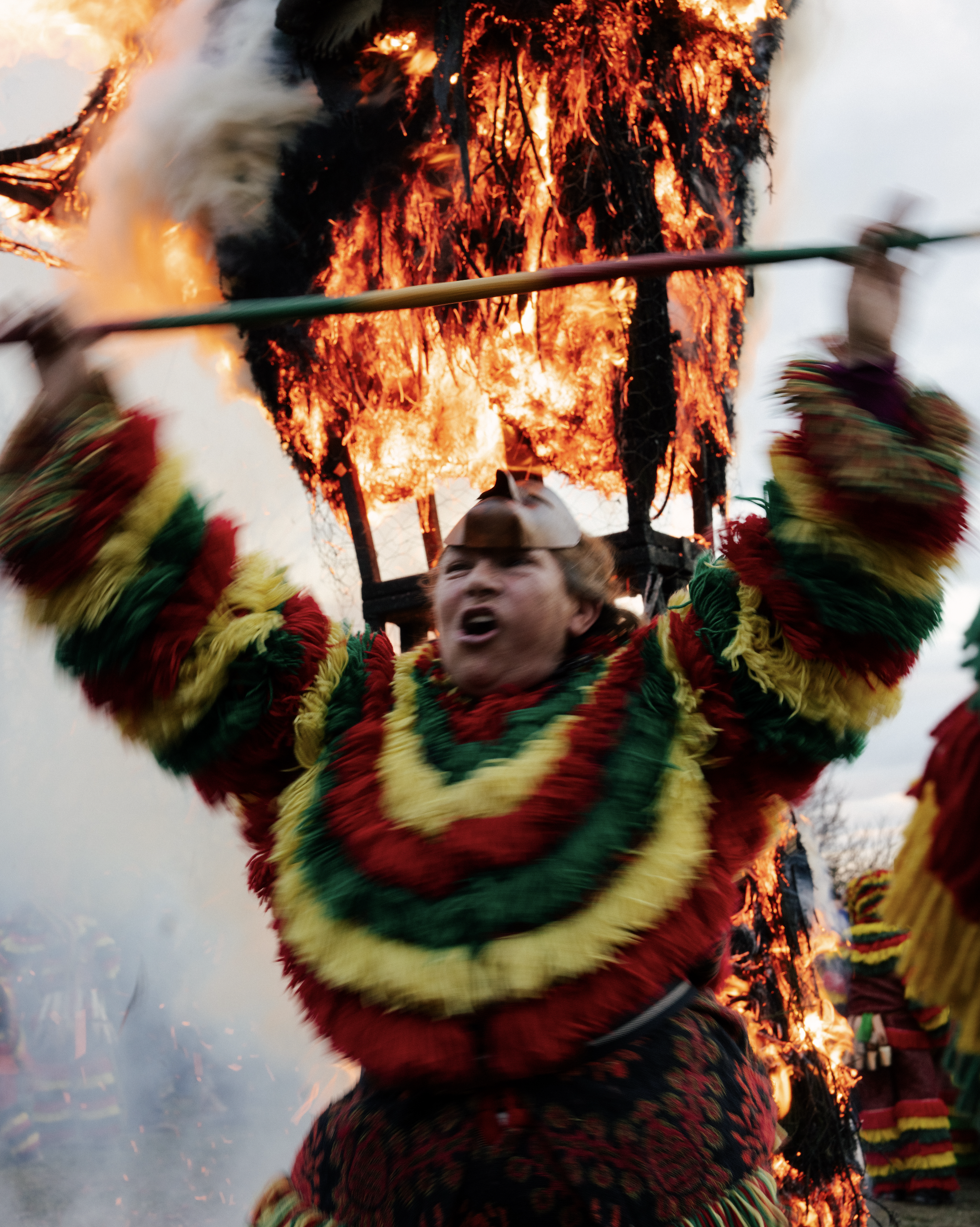 Person dressed in colorful traditional costume participating in a fire festival, holding a flaming torch with a background of burning wood and sparks.