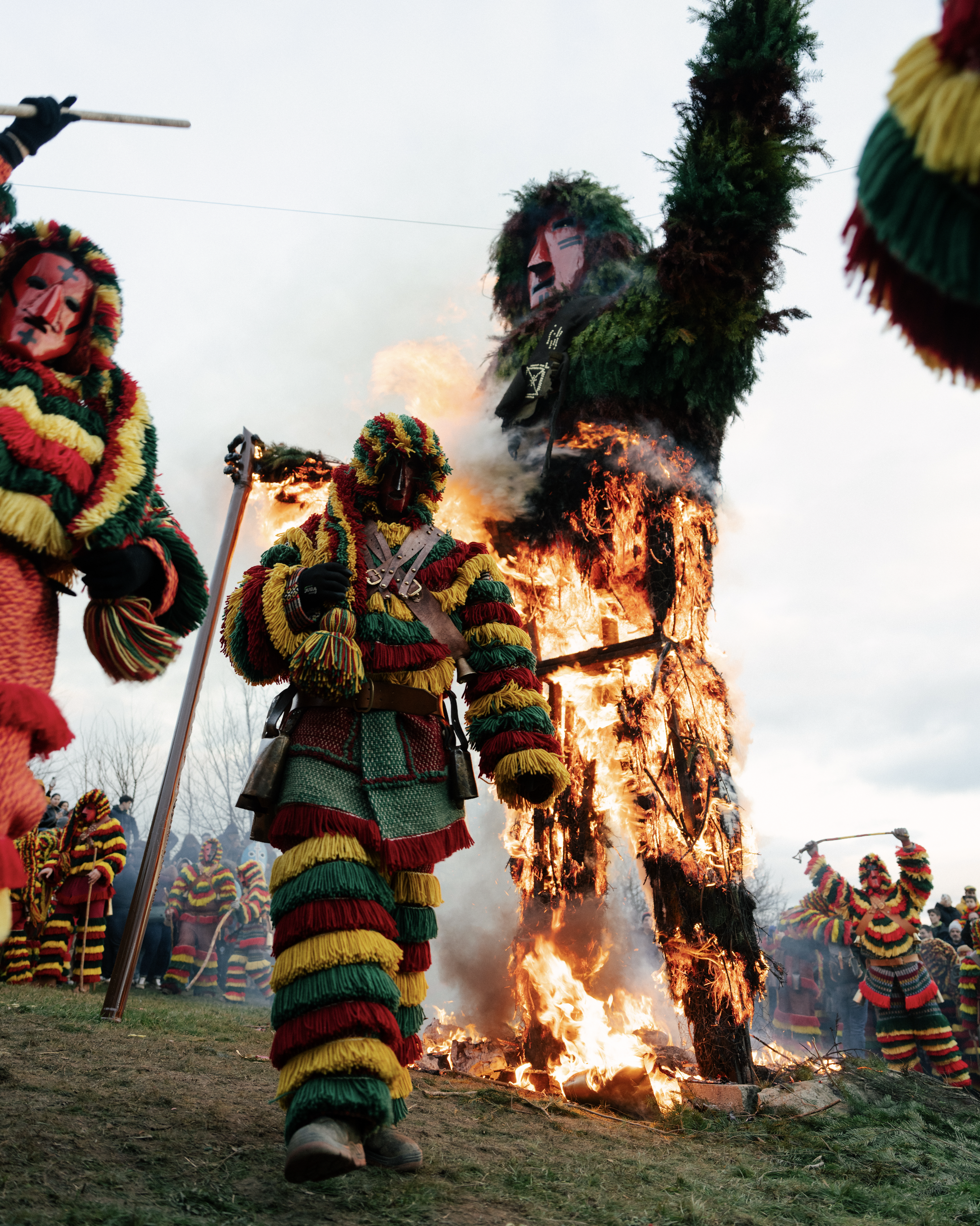Participants in colorful traditional costumes burning a large wooden effigy during a cultural festival, with onlookers in the background.