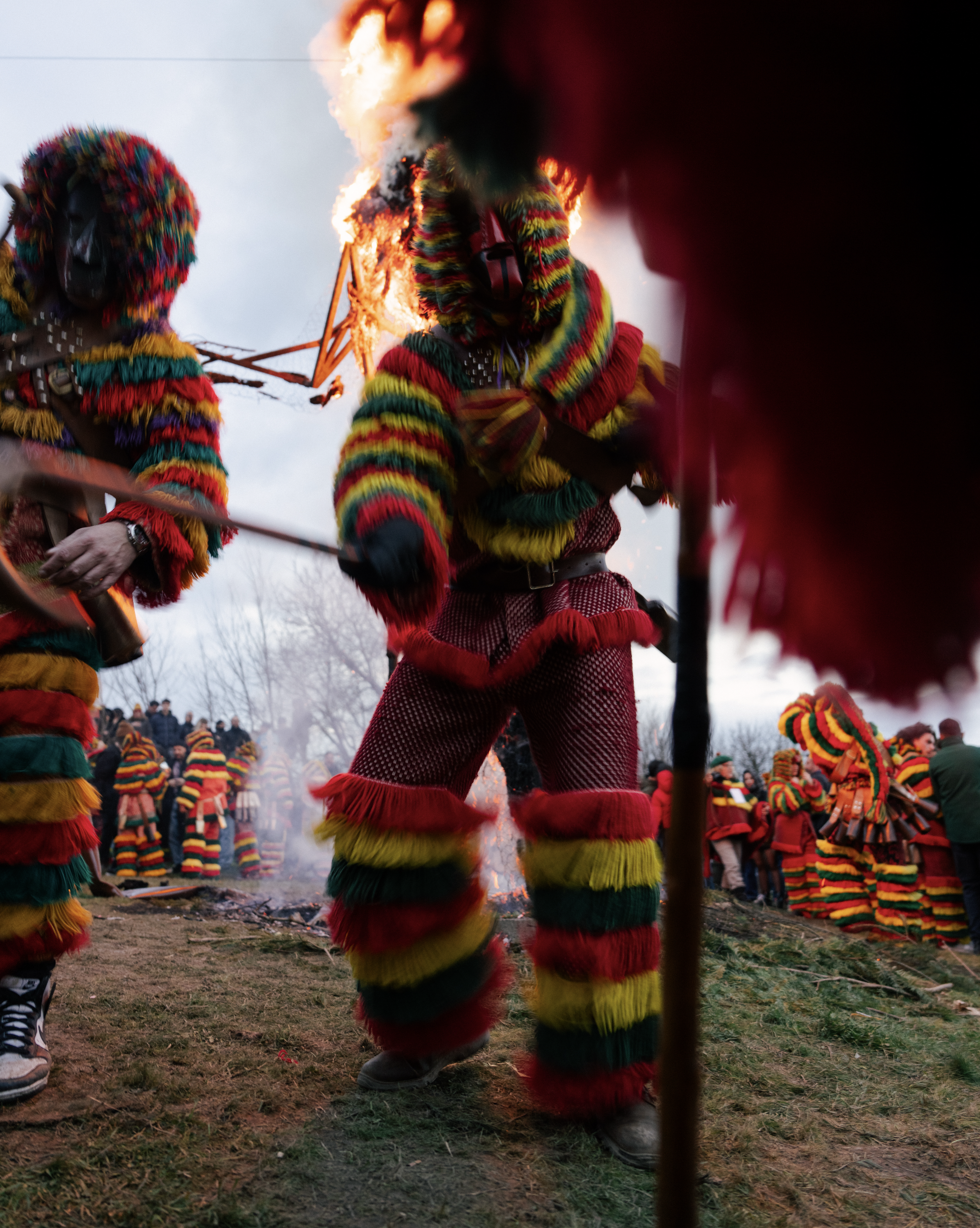 People dressed in colorful, furry costumes with masks, participating in a traditional celebration around a fire outdoors on a cloudy day.