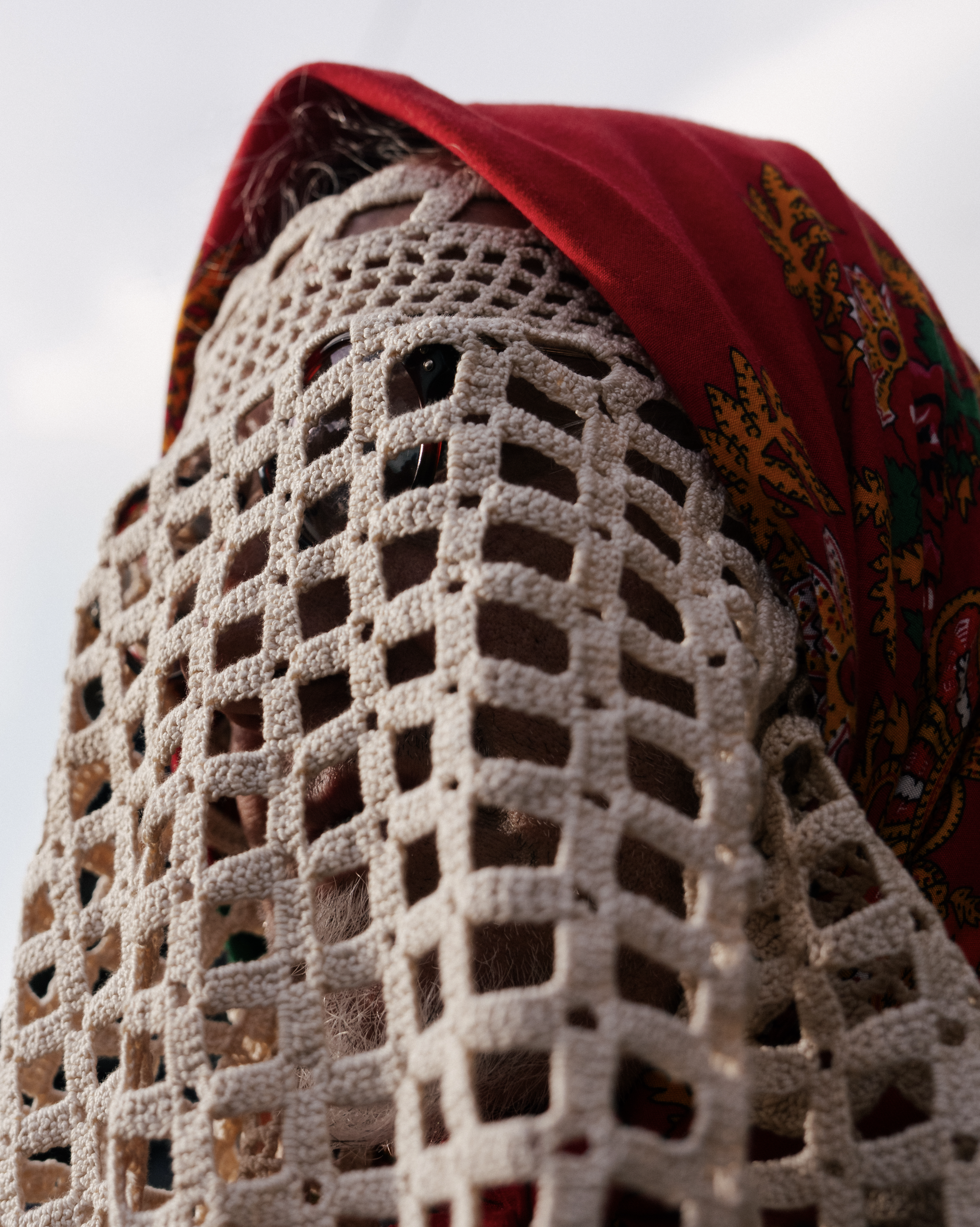 Close-up of a person wearing a crochet mask and a red bandana with colorful patterns over their head, against a cloudy sky.