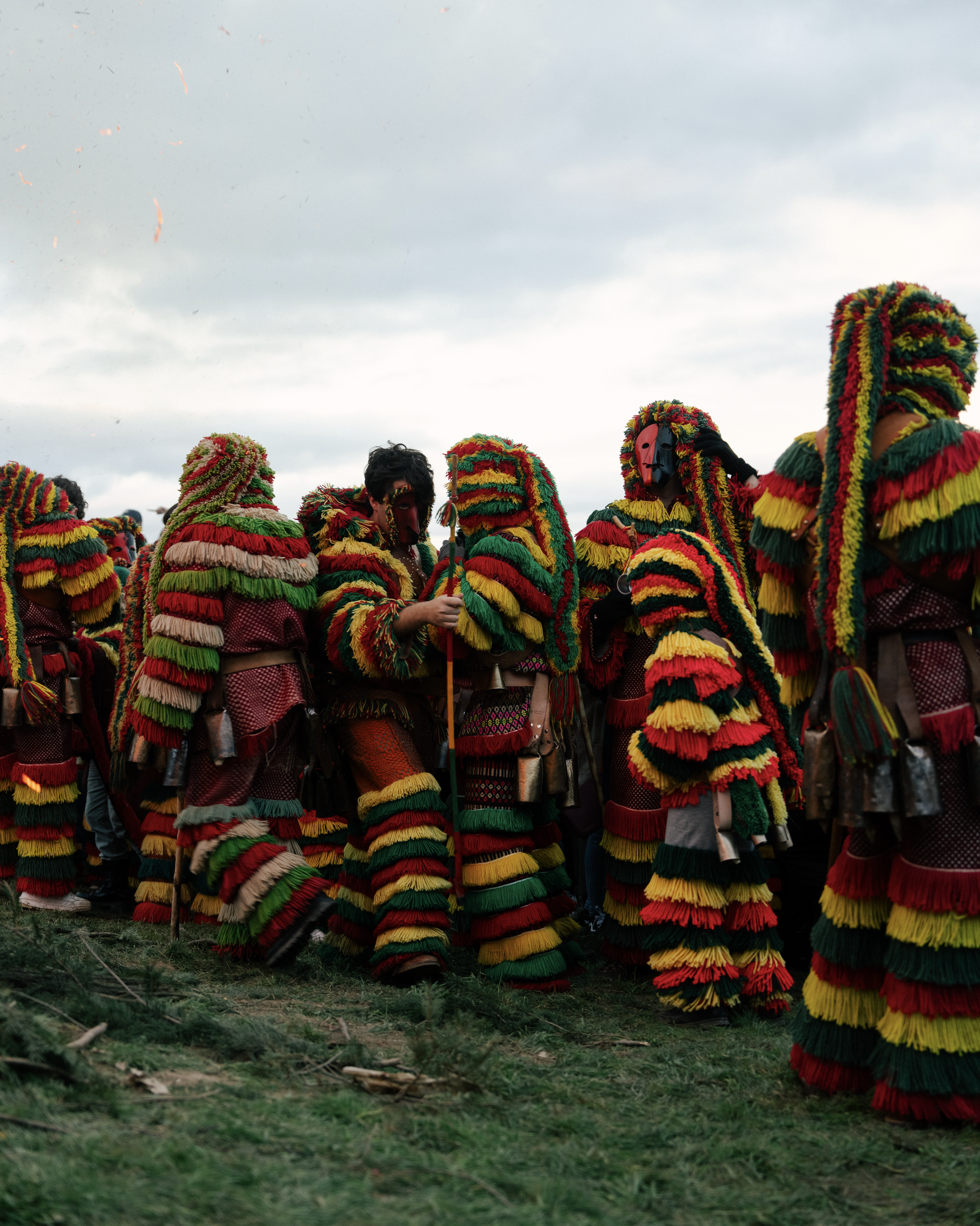 A group of people dressed in colorful traditional costumes with masks, participating in a cultural festival outdoors. The costumes are layered with multicolored fringes, primarily red, yellow, green, and black, and the participants are standing on grassy ground under a cloudy sky.
