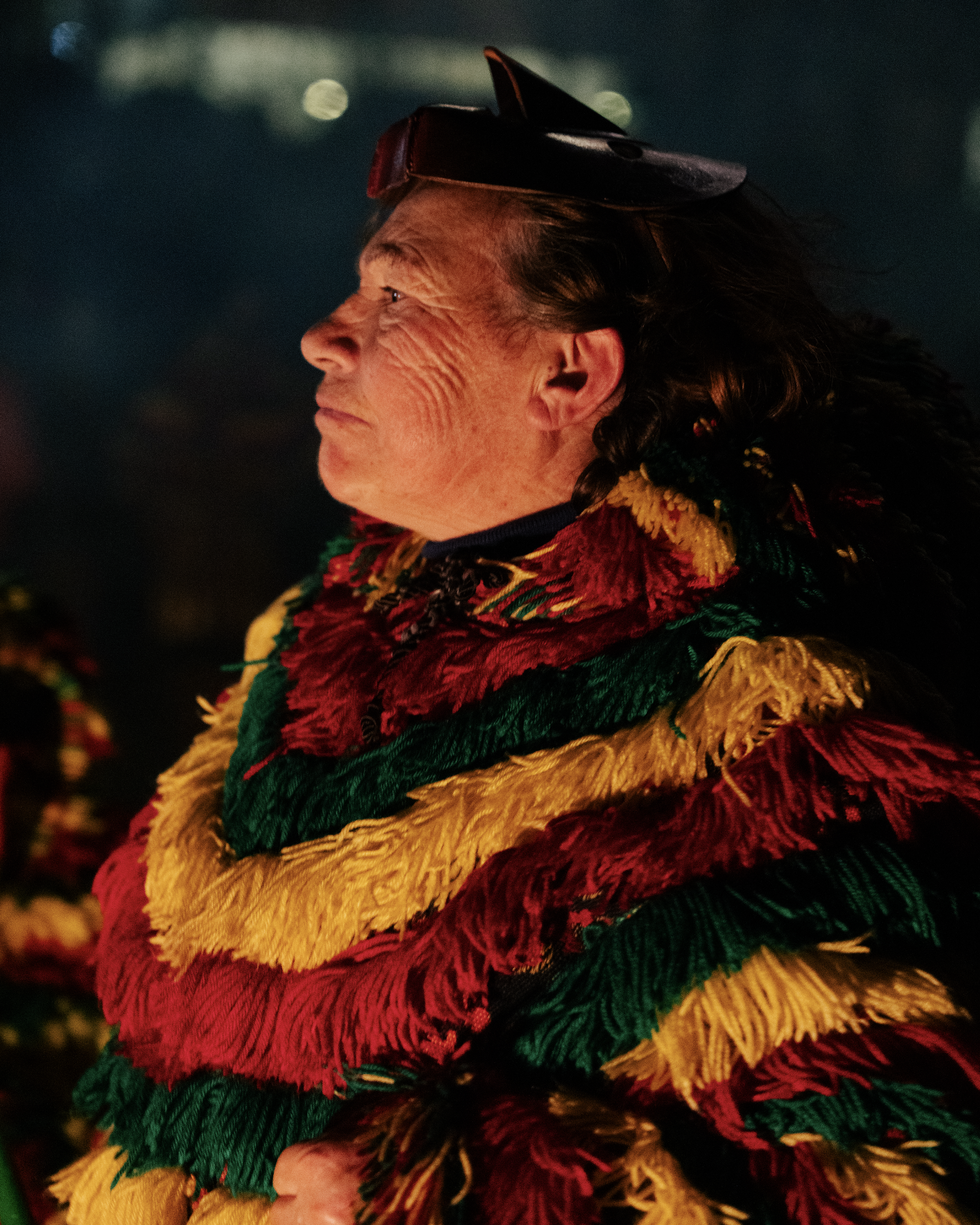 Close-up side profile of an elderly woman with light skin, wearing a colorful festive scarf and a small black hat, with a background of blurred lights.
