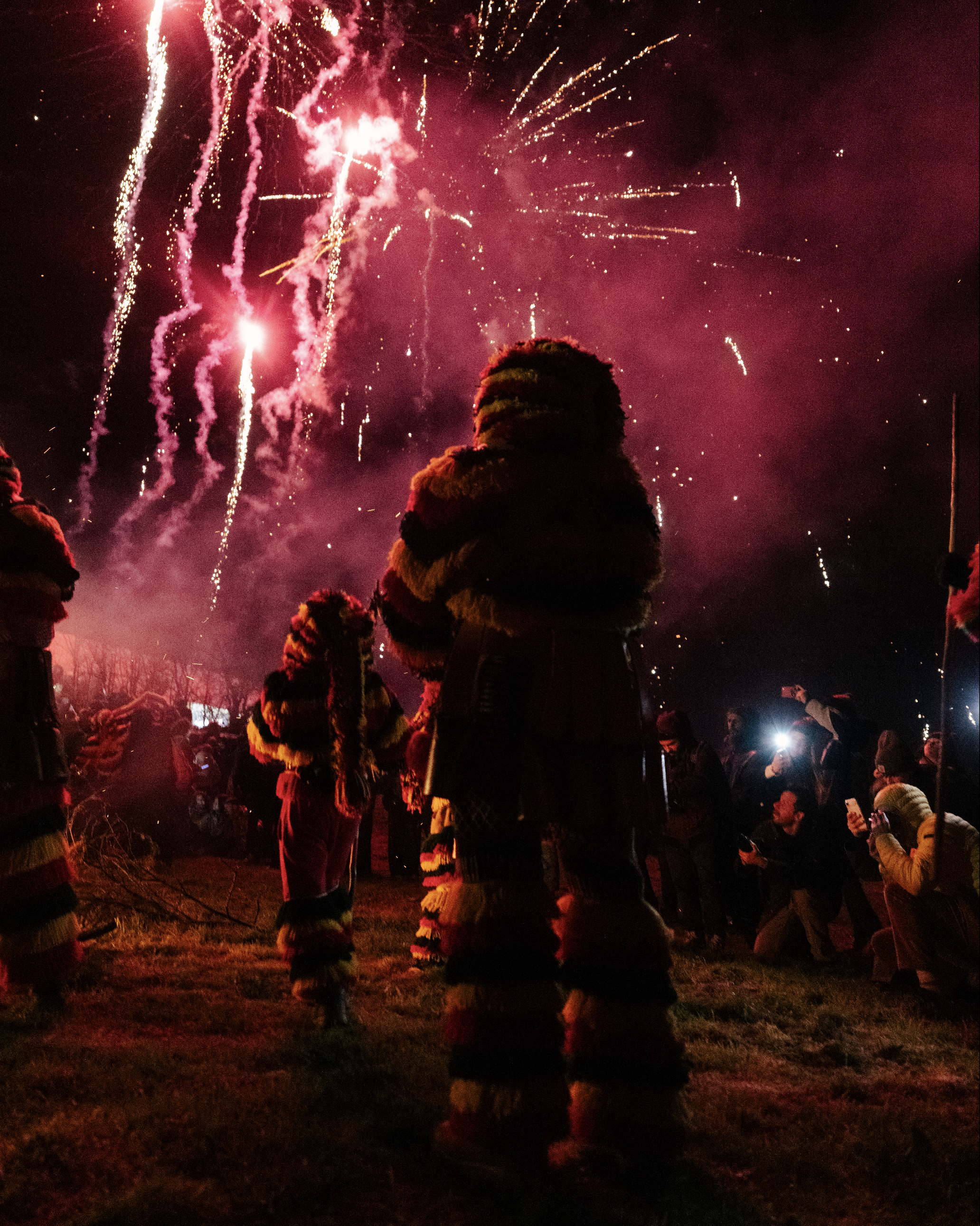 People gathered outdoors at night watching fireworks display with red and gold explosions in the sky, some individuals are recording or taking pictures with their phones.