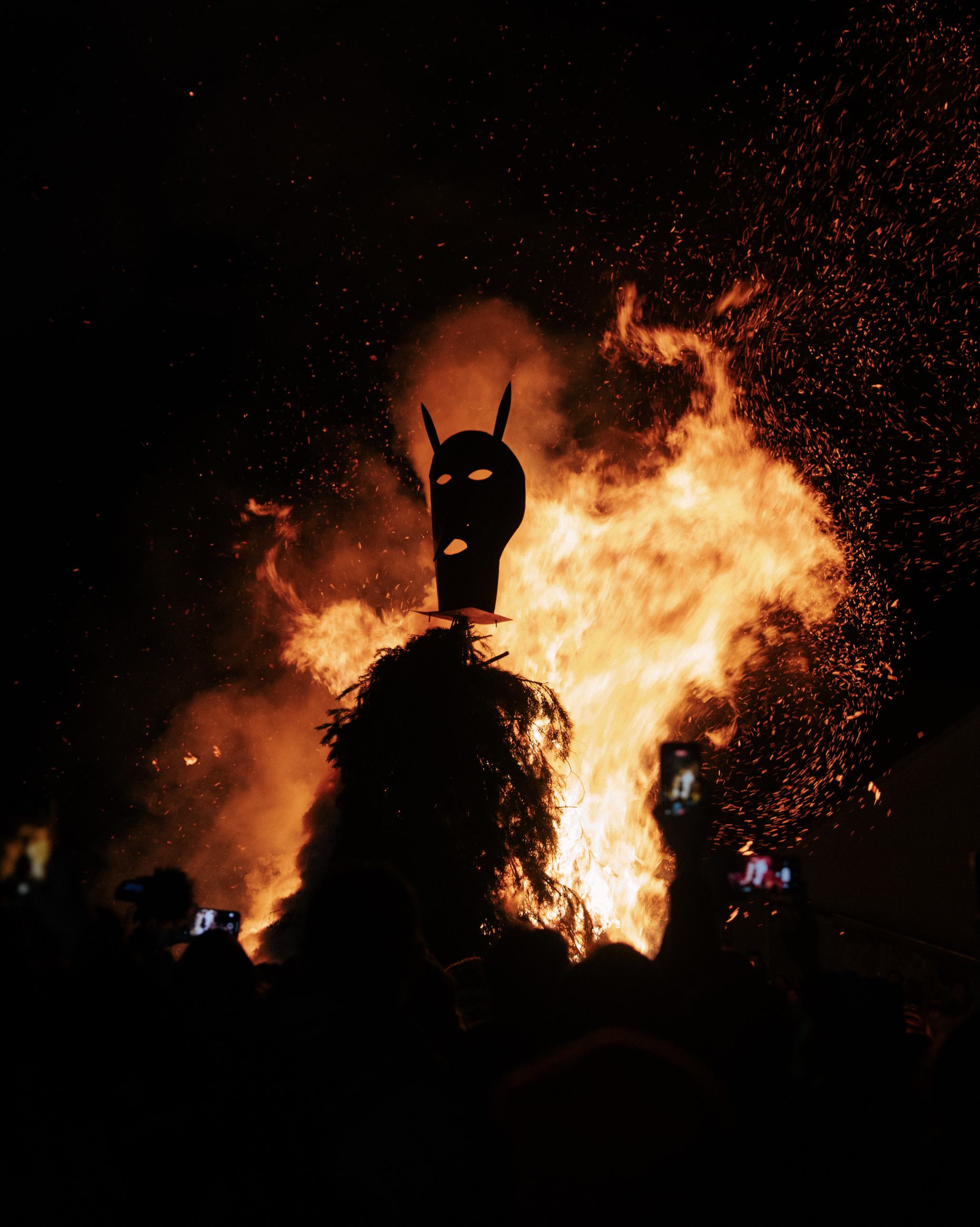 Large fire with flames and sparks rising into the night sky, with a black mask-like figure with bunny ears in front of the fire, and silhouettes of people taking photos or videos at the bottom.