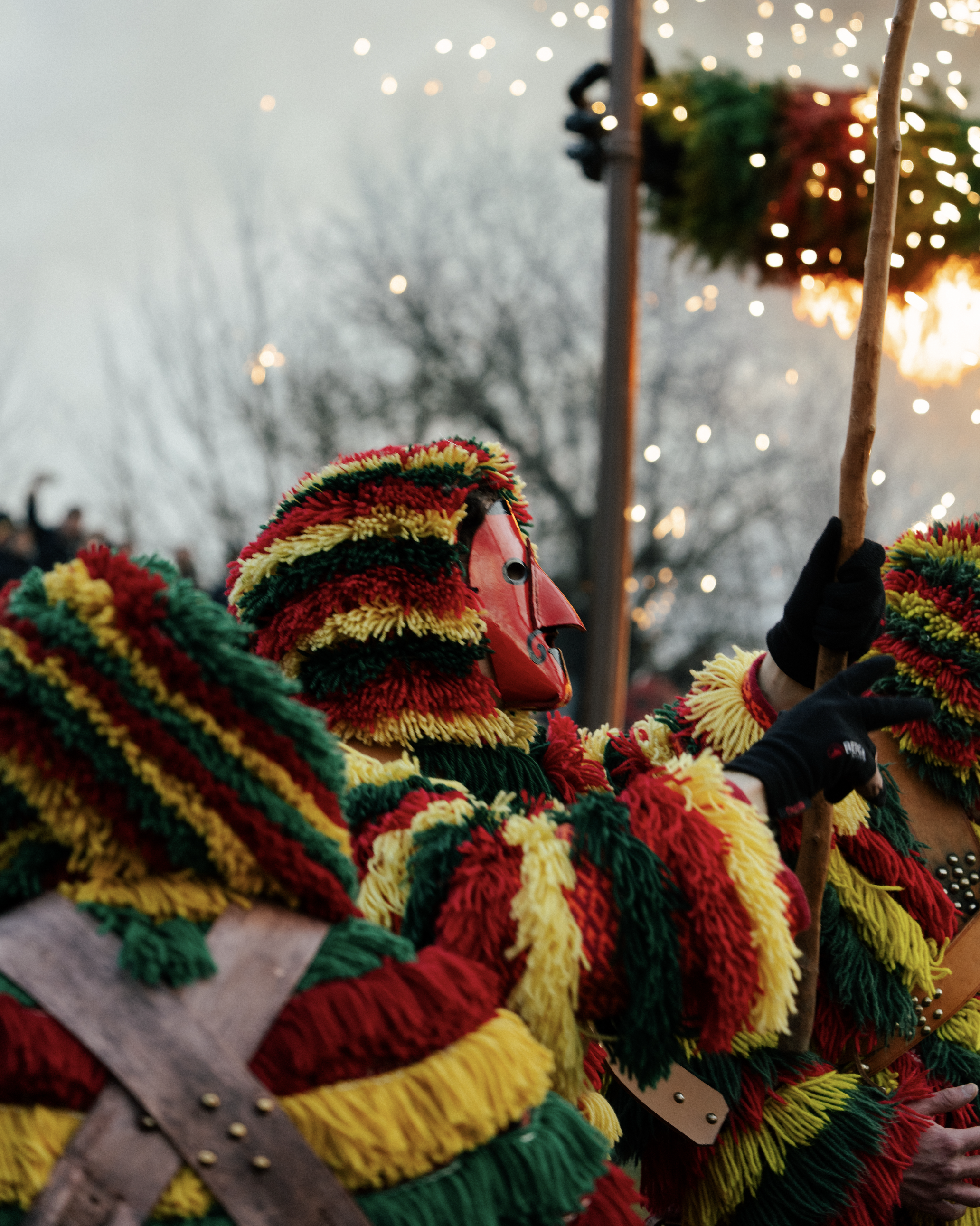 Close-up of colorful woven hats with red, green, yellow, and black yarn, displayed for sale at an outdoor market, with blurred trees and string lights in the background.