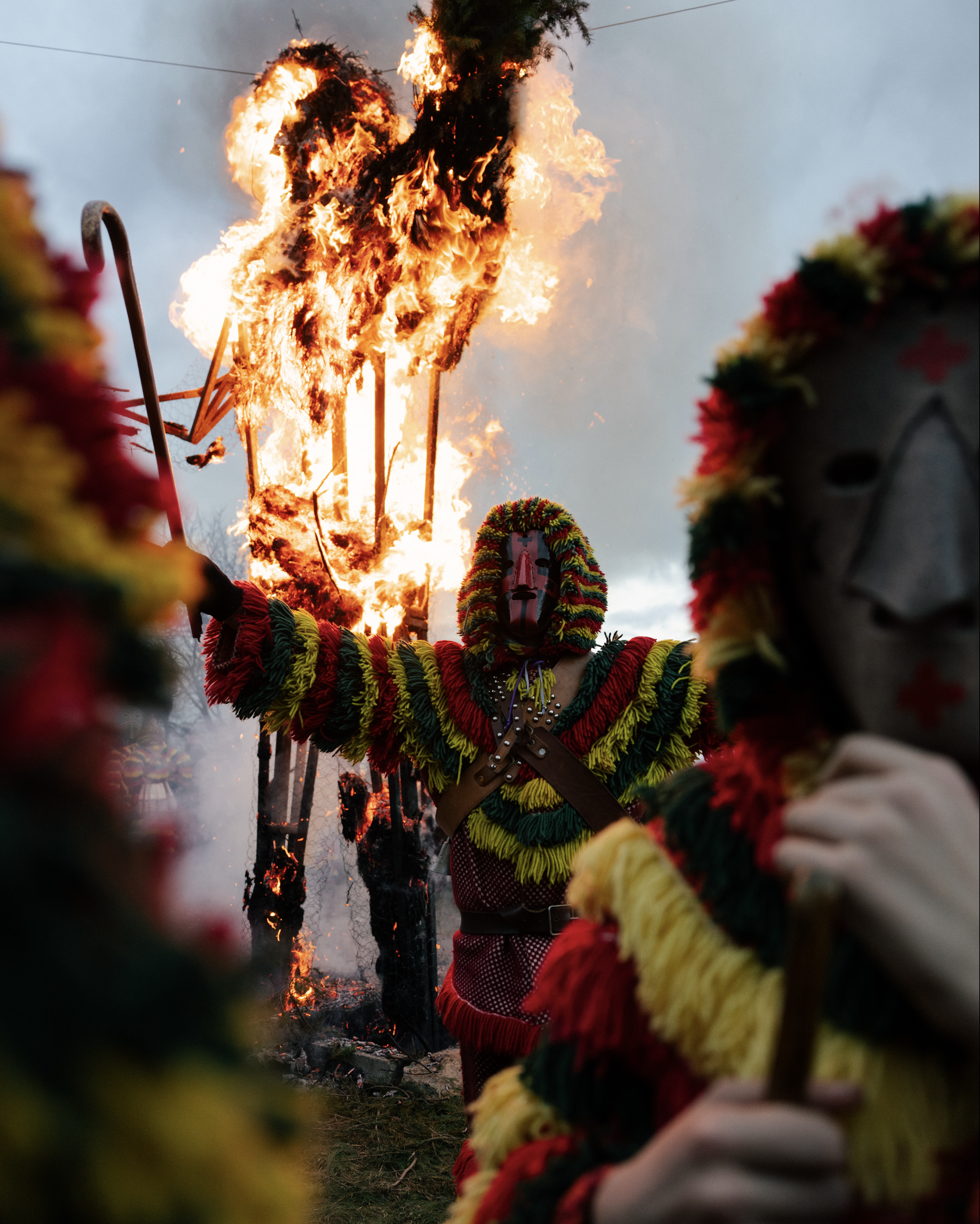 A person in traditional colorful attire and a mask participating in a cultural festival or ritual, with a large fire burning in the background, possibly during a ceremony or celebration.