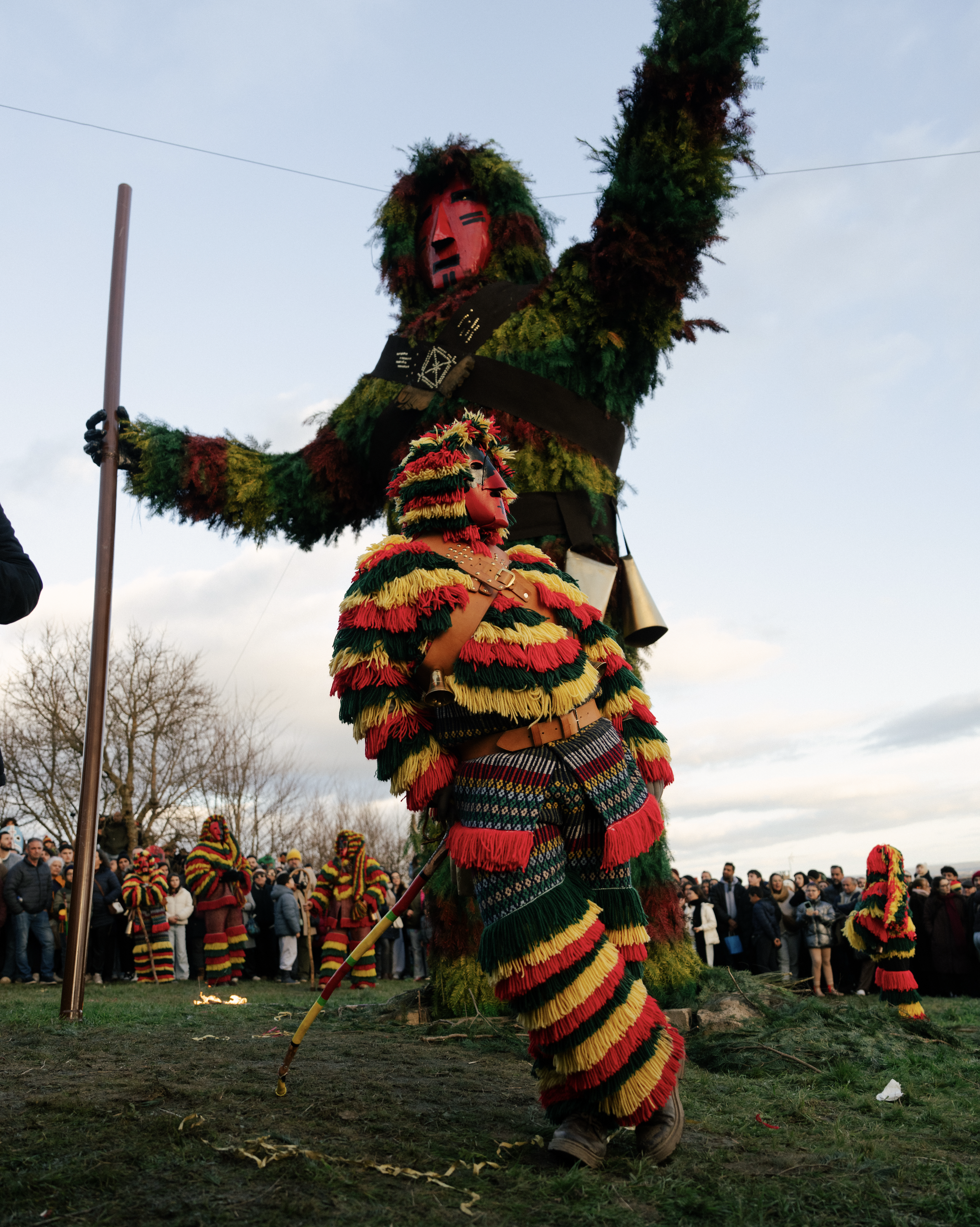 A person dressed in a colorful traditional costume with a large mask on their face, participating in a cultural festival or parade. The costume includes multicolored fringe and the person is holding a long pole. There are onlookers in the background, and the scene appears to be outdoors during daytime with a cloudy sky.