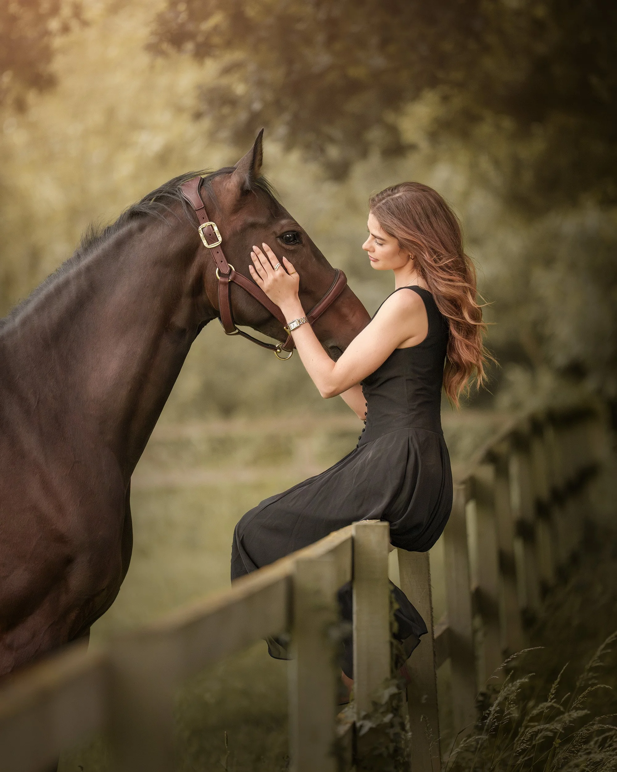 A woman with long, wavy red hair wearing a black dress gently touching the face of a brown horse through a wooden fence in an outdoor setting with a blurred background of trees and grass.