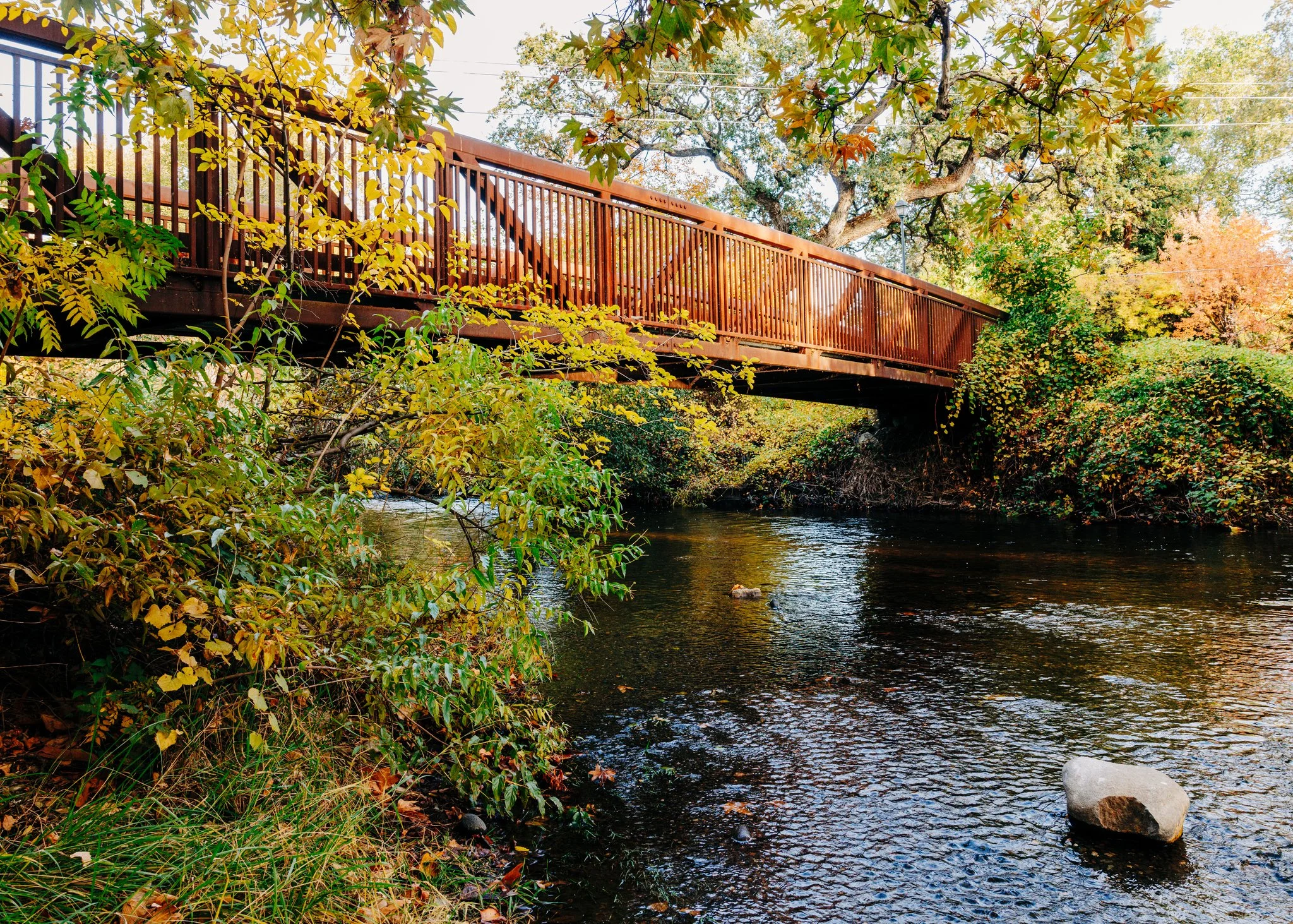 A wooden bridge spanning over a small river, surrounded by trees with green and orange autumn leaves.