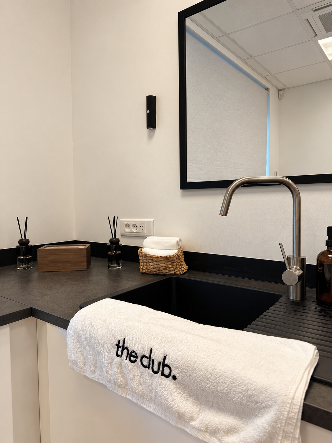Un lavabo avec un savon en spray, un miroir, des brosses à cheveux, et des bocaux sur un comptoir, avec une serviette suspendue portant le texte 'the club'.