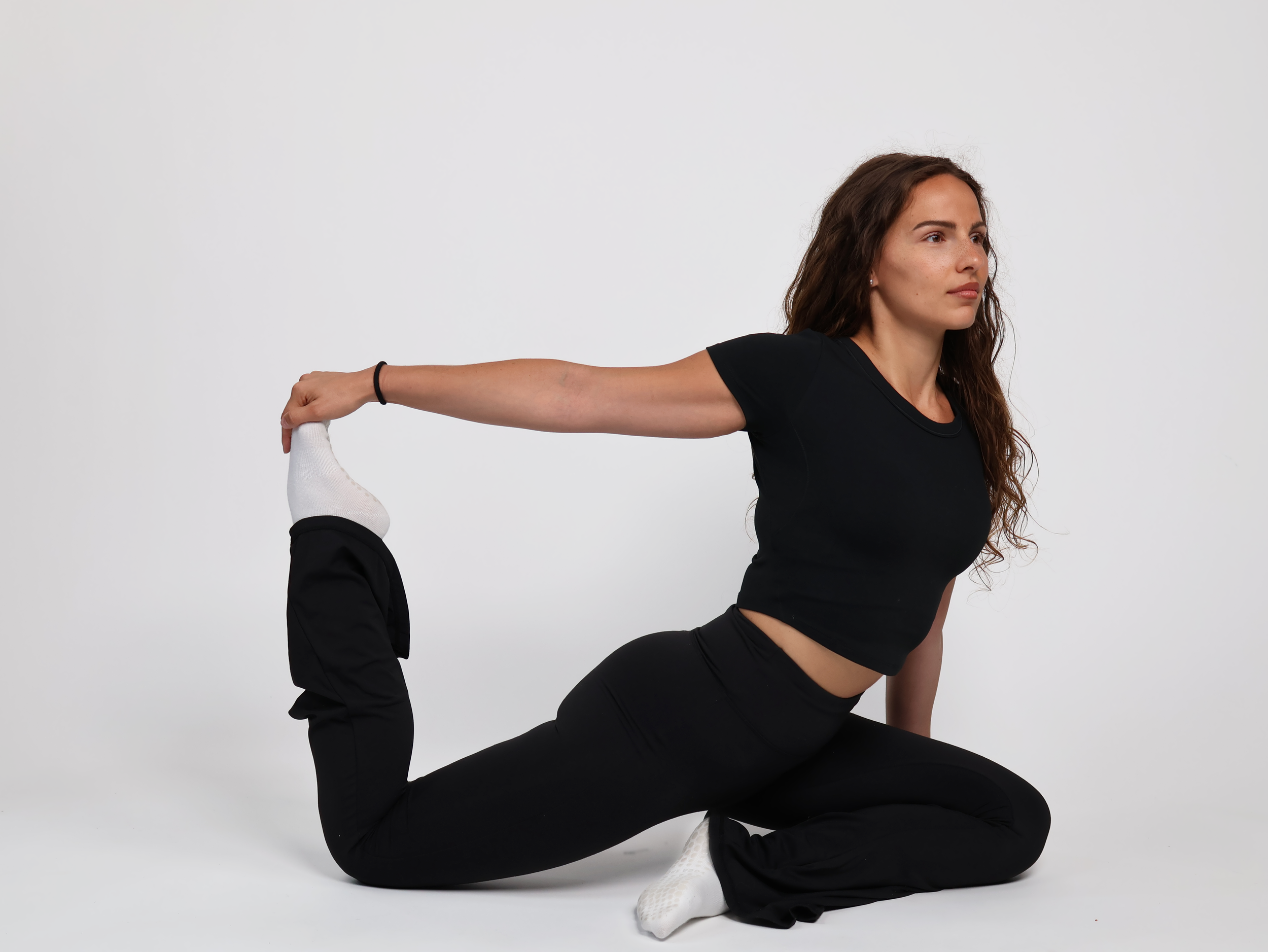 Femme faisant une posture de yoga en position de fente arrière avec une main tenant le pied derrière elle, dans un studio au fond blanc.