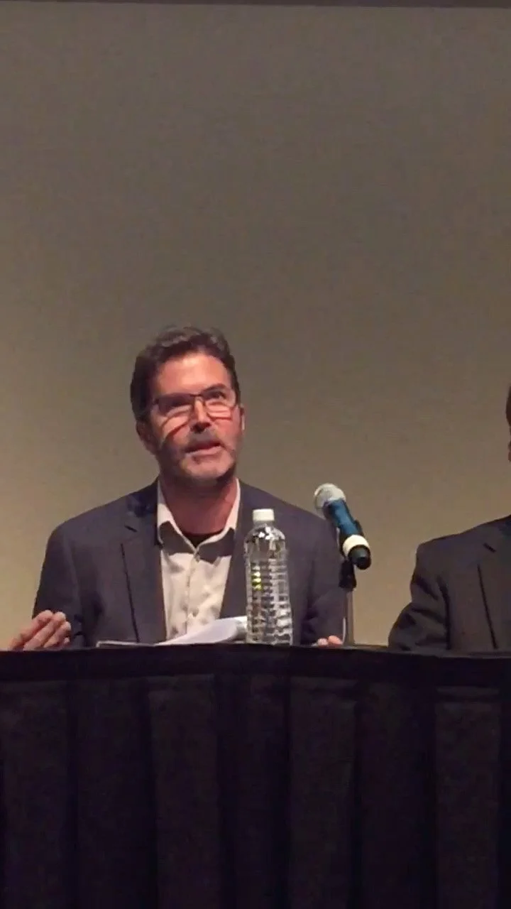 A man wearing glasses, a suit, and a white shirt sitting at a table with a bottle of water and a microphone, speaking at a panel or conference.