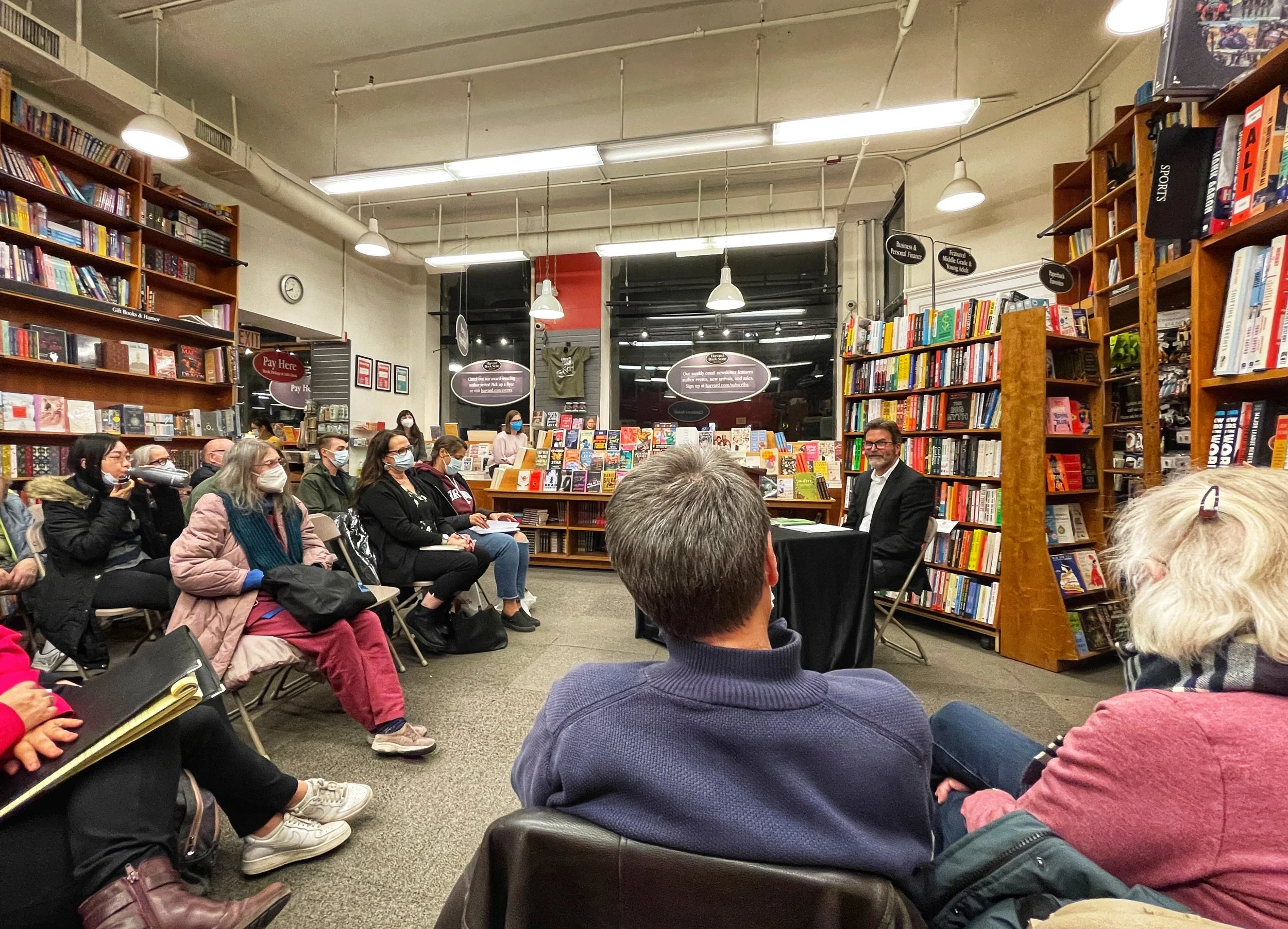 A book signing event inside a bookstore featuring a seated author at a table and an audience of around twenty people, all wearing face masks, listening attentively.