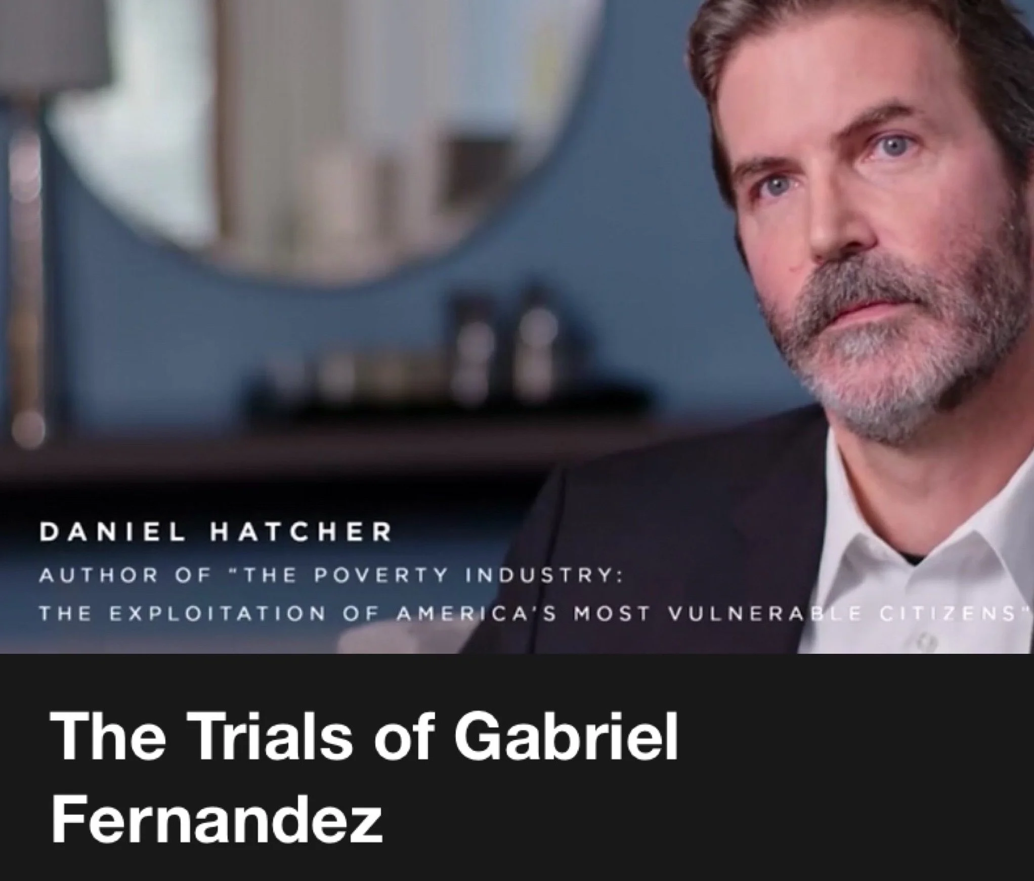 Close-up of Daniel L. Hatcher in Netflix interview, with beard and brown hair, wearing a black suit and white shirt, sitting in a room with a blurred background and a blue wall in the background.