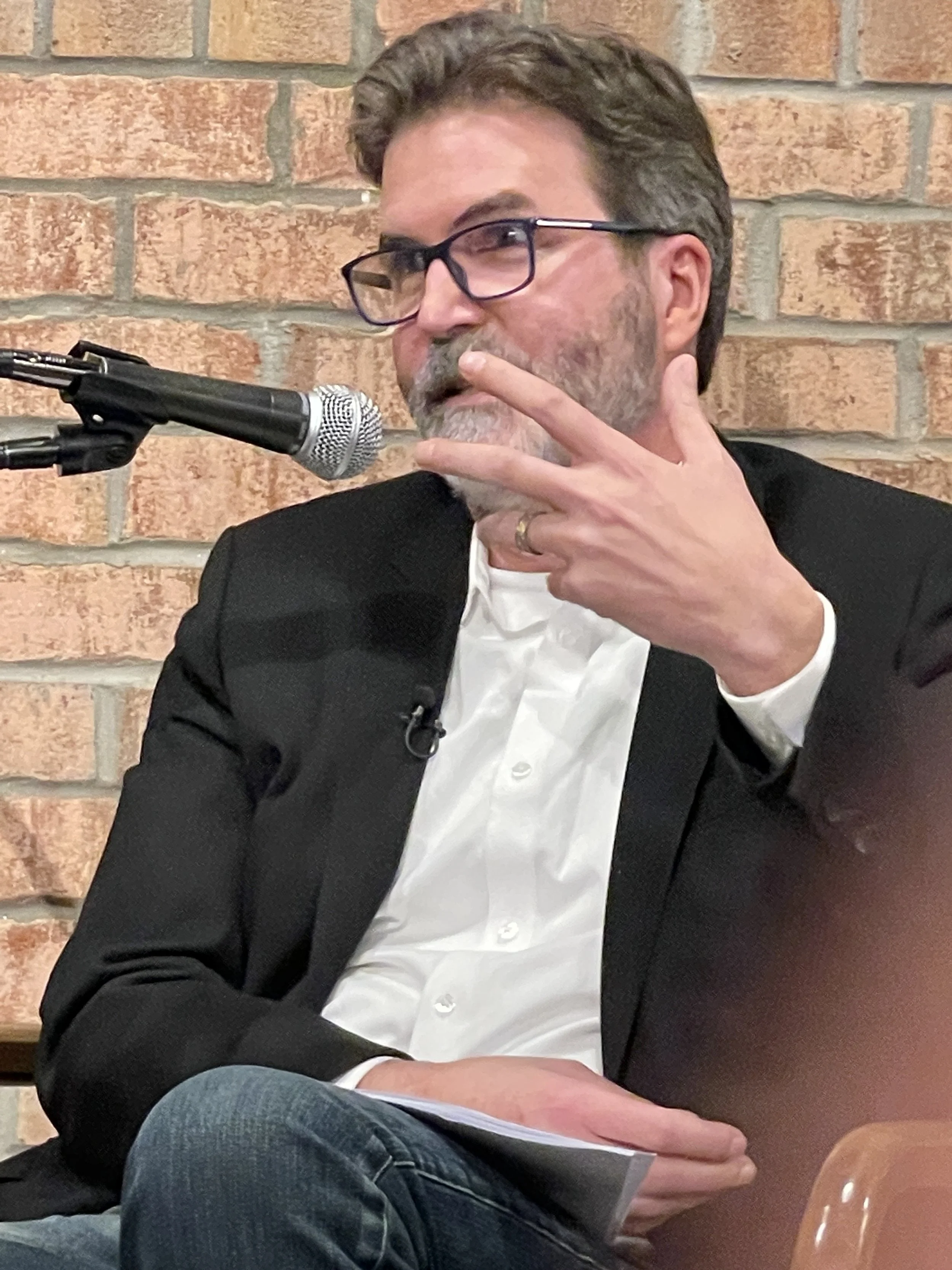 A man with glasses and a beard, wearing a black blazer and white shirt, is speaking into a microphone while holding some papers in his lap. He is sitting in front of a brick wall.