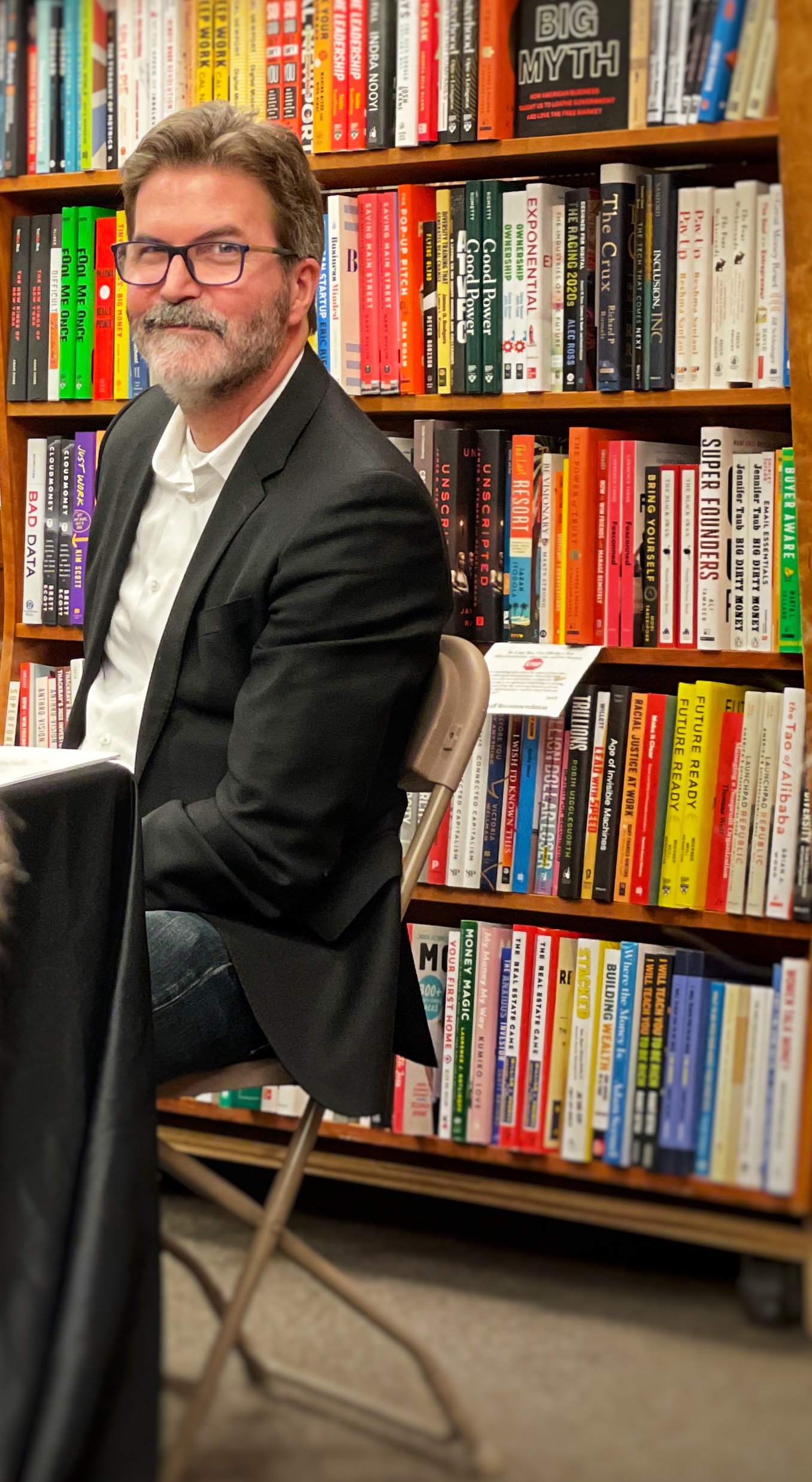 A man with glasses and a beard, dressed in a black blazer and white shirt, sitting in a library with bookshelves filled with colorful books in the background.