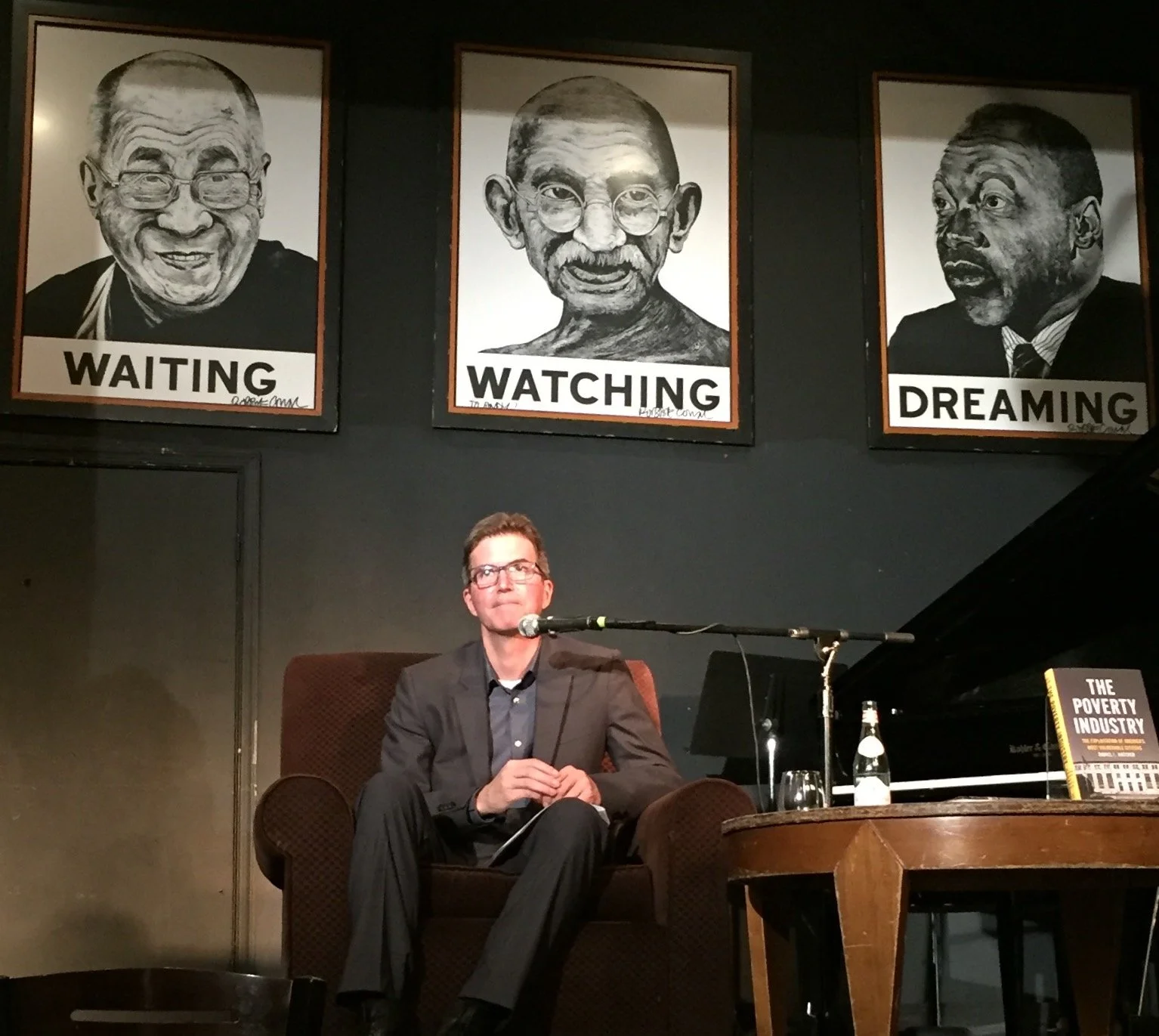 A man in a suit sits in a brown armchair on a stage, holding a notepad and sitting in front of a microphone. Behind him are three portraits with labels "Waiting," "Watching," and "Dreaming," depicting caricatures of prominent figures.