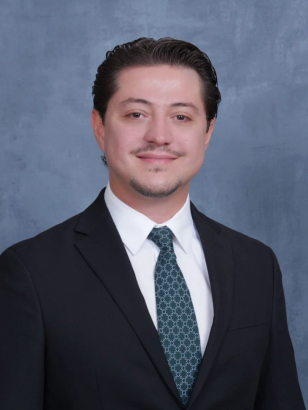 Headshot of a man with dark hair, wearing a black suit, white shirt, patterned tie, and smiling at the camera against a blue background.