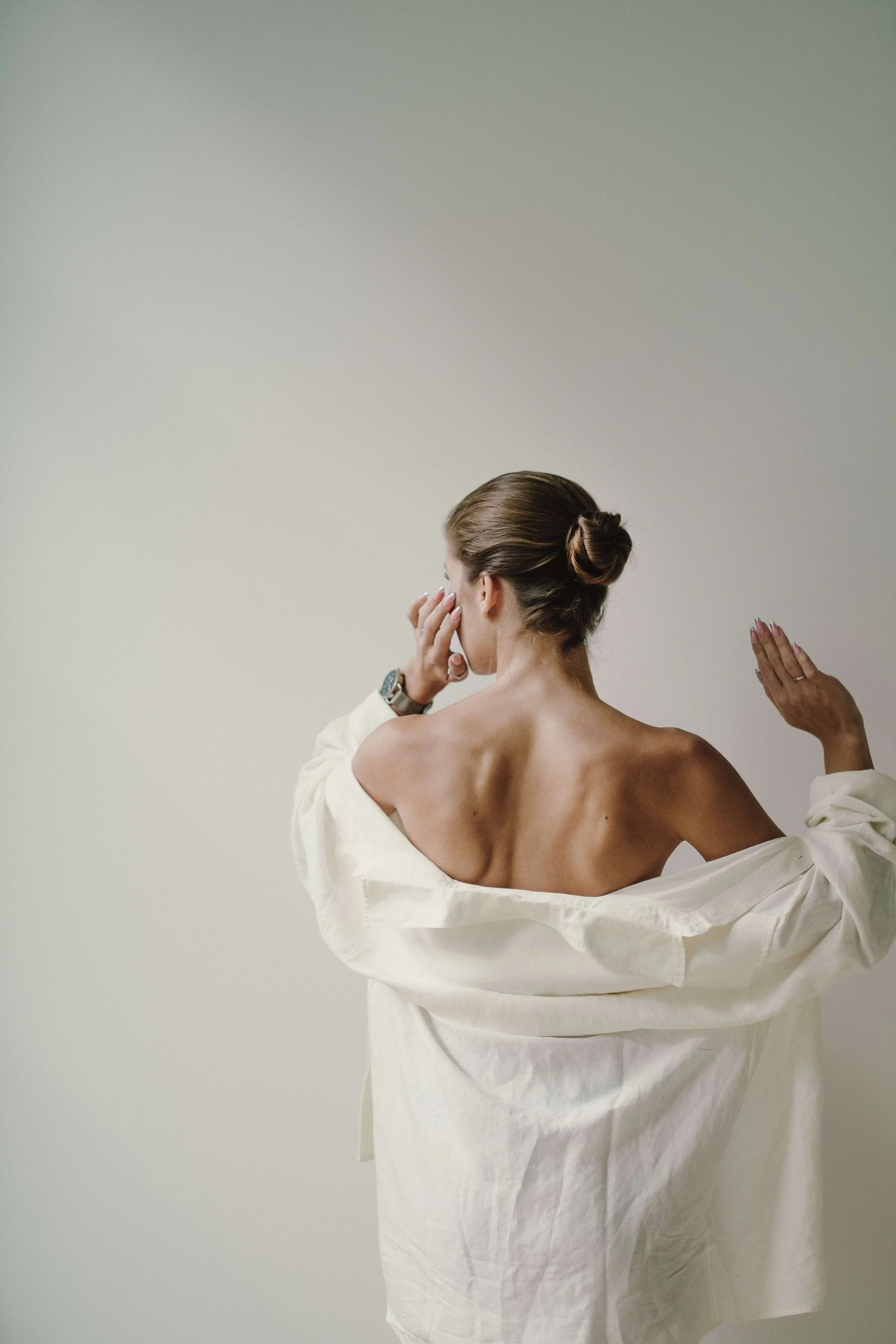 Back view of a woman with brown hair styled in a bun, wearing a loose white shirt, holding her hand to her face, with another person reaching towards her shoulder.