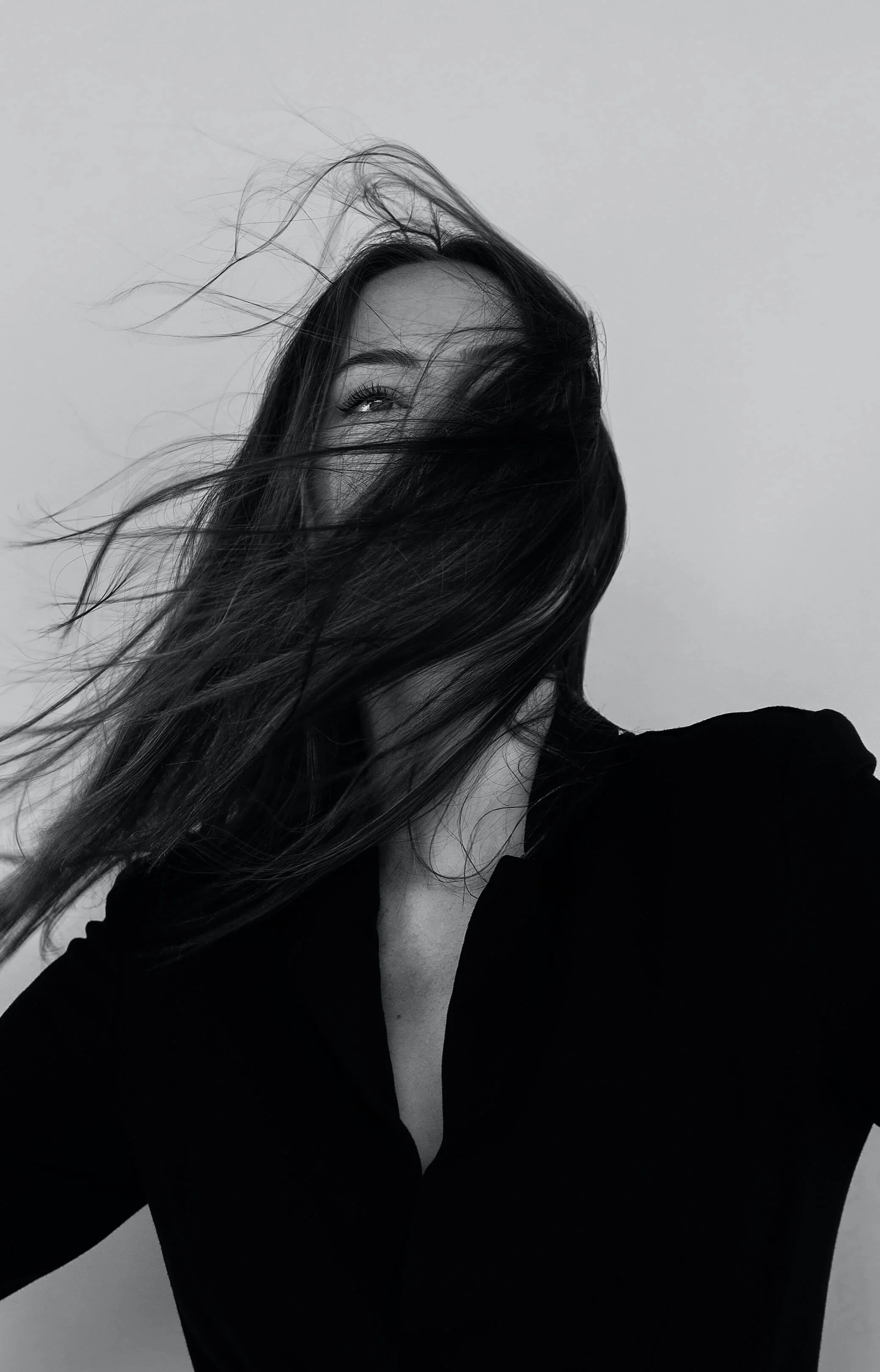 Black and white photograph of a woman with long hair, partially covering her face, wearing a black top, with a plain background.