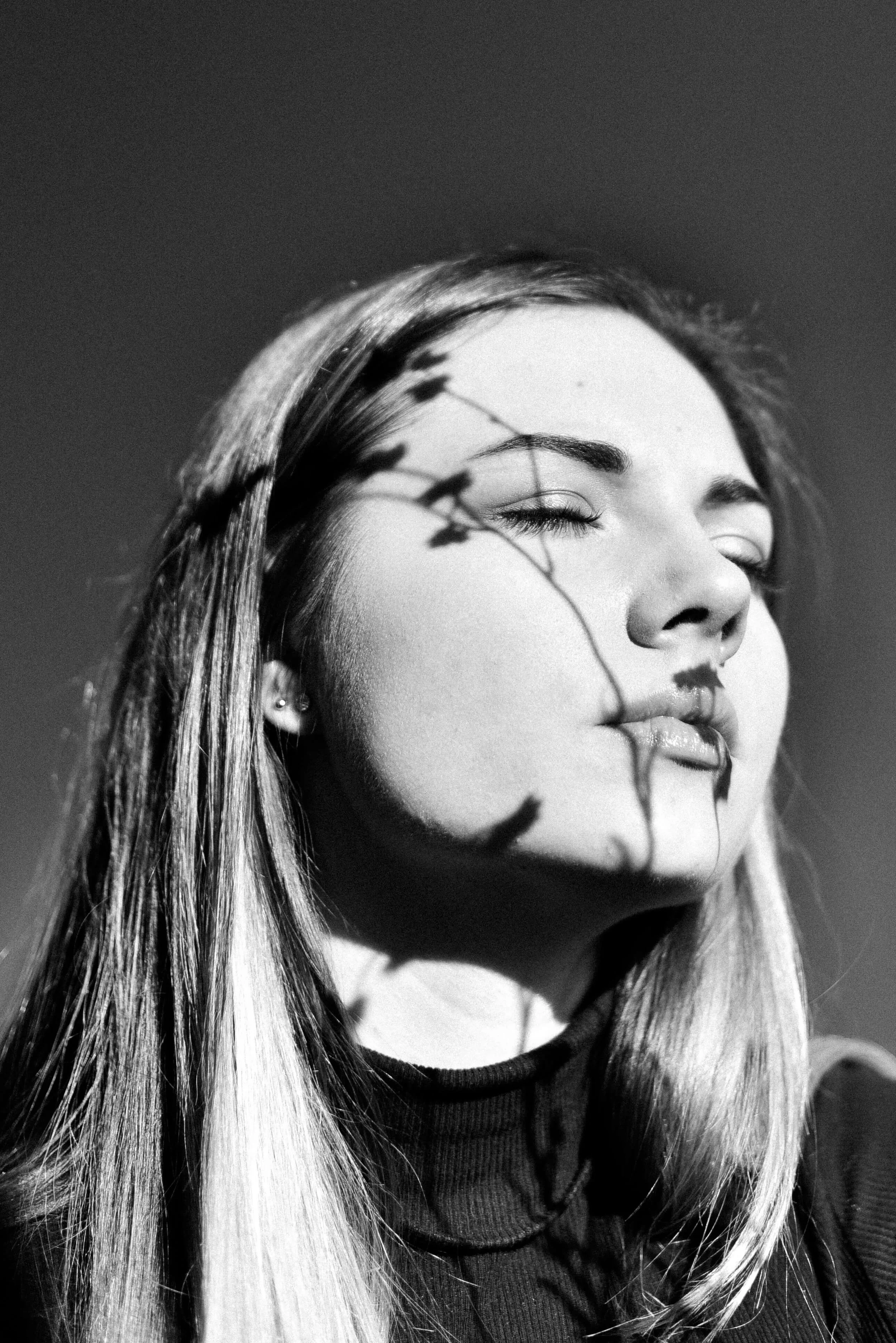 A black-and-white close-up photo of a woman with her eyes closed, her face slightly tilted, with a shadow of a plant cast across her face. She has long hair, earrings, and is wearing a dark top.