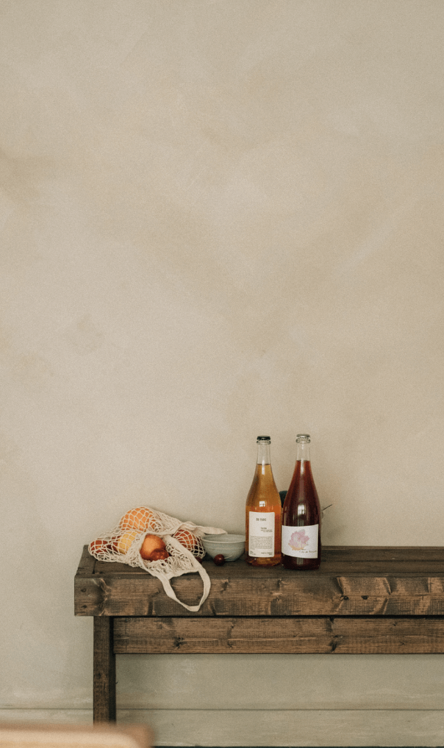 A wooden table against a plain wall holding two bottles of drinks, a bowl, and a reusable net bag with onions.