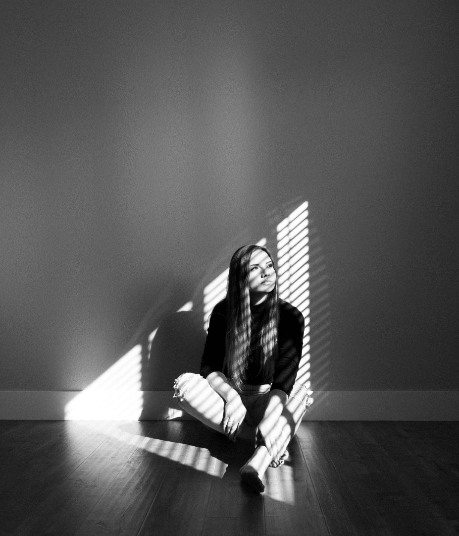 A young woman with long hair sits cross-legged on the floor next to a plain wall, with sunlight filtering through blinds creating striped shadows on her and the wall.