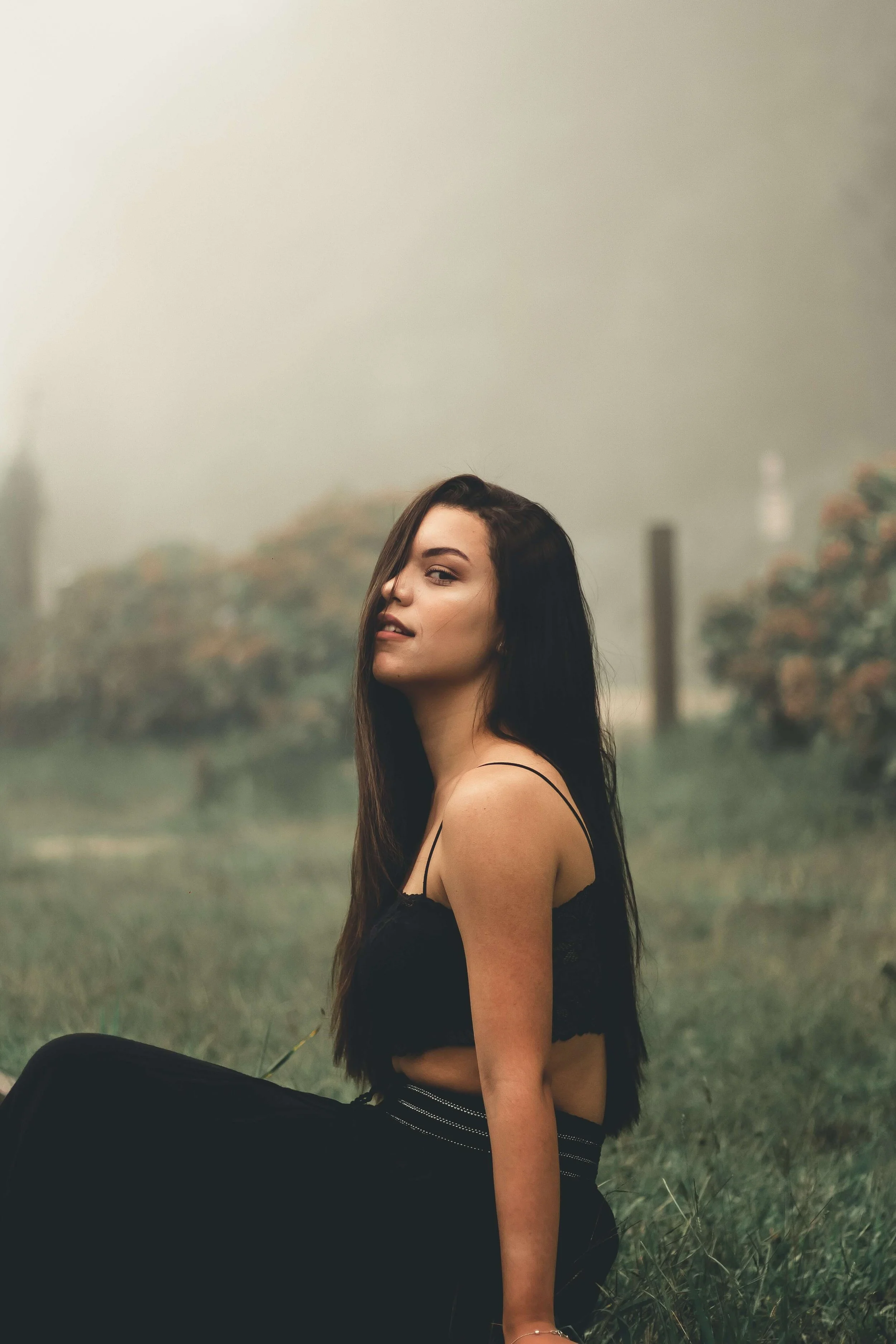 A young woman with long dark hair sitting outdoors on grass, wearing a black top and pants, with a hazy, cloudy sky and trees in the background.