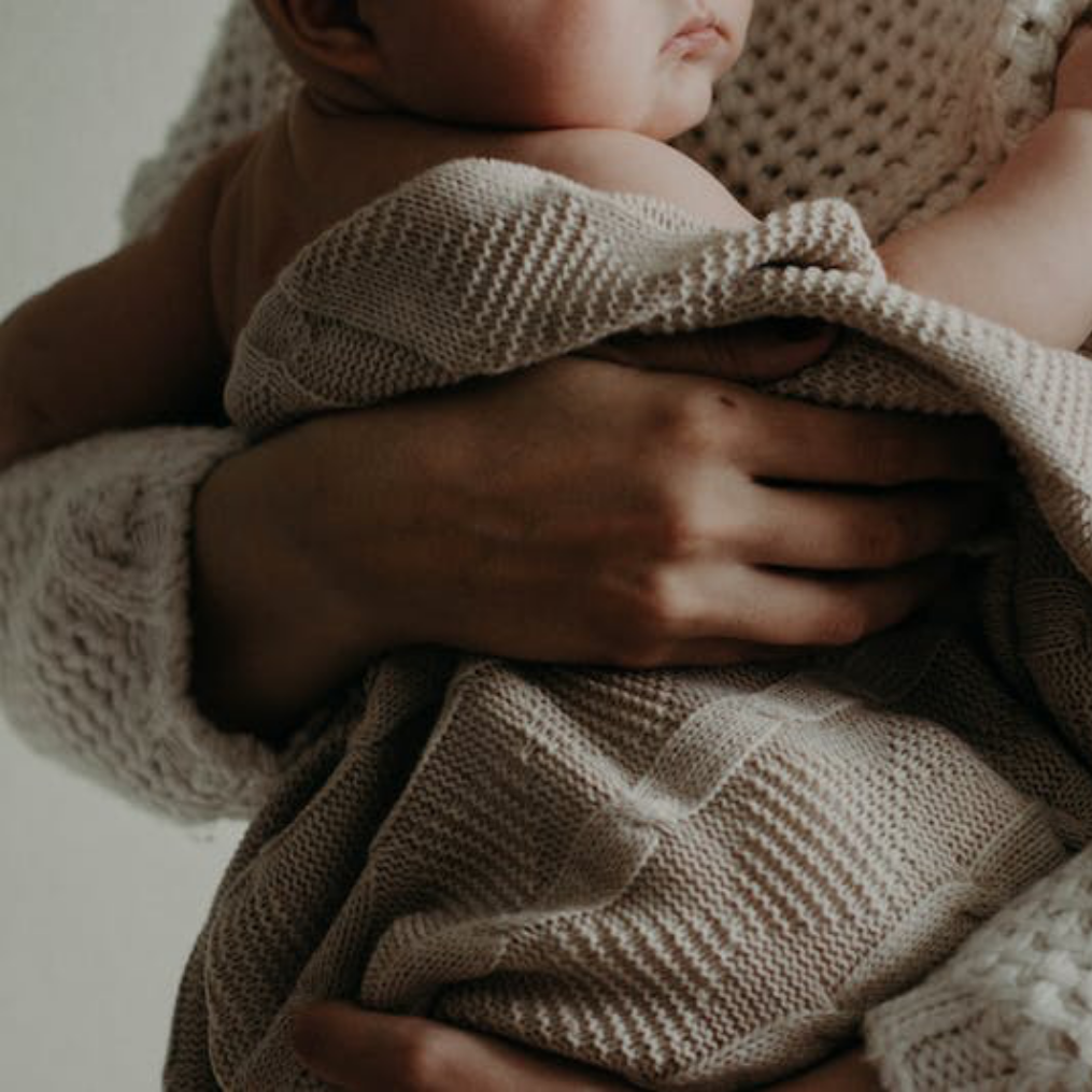 Close-up of a person holding a baby wrapped in a beige knitted blanket, focusing on the person's hand gently supporting the baby's head.