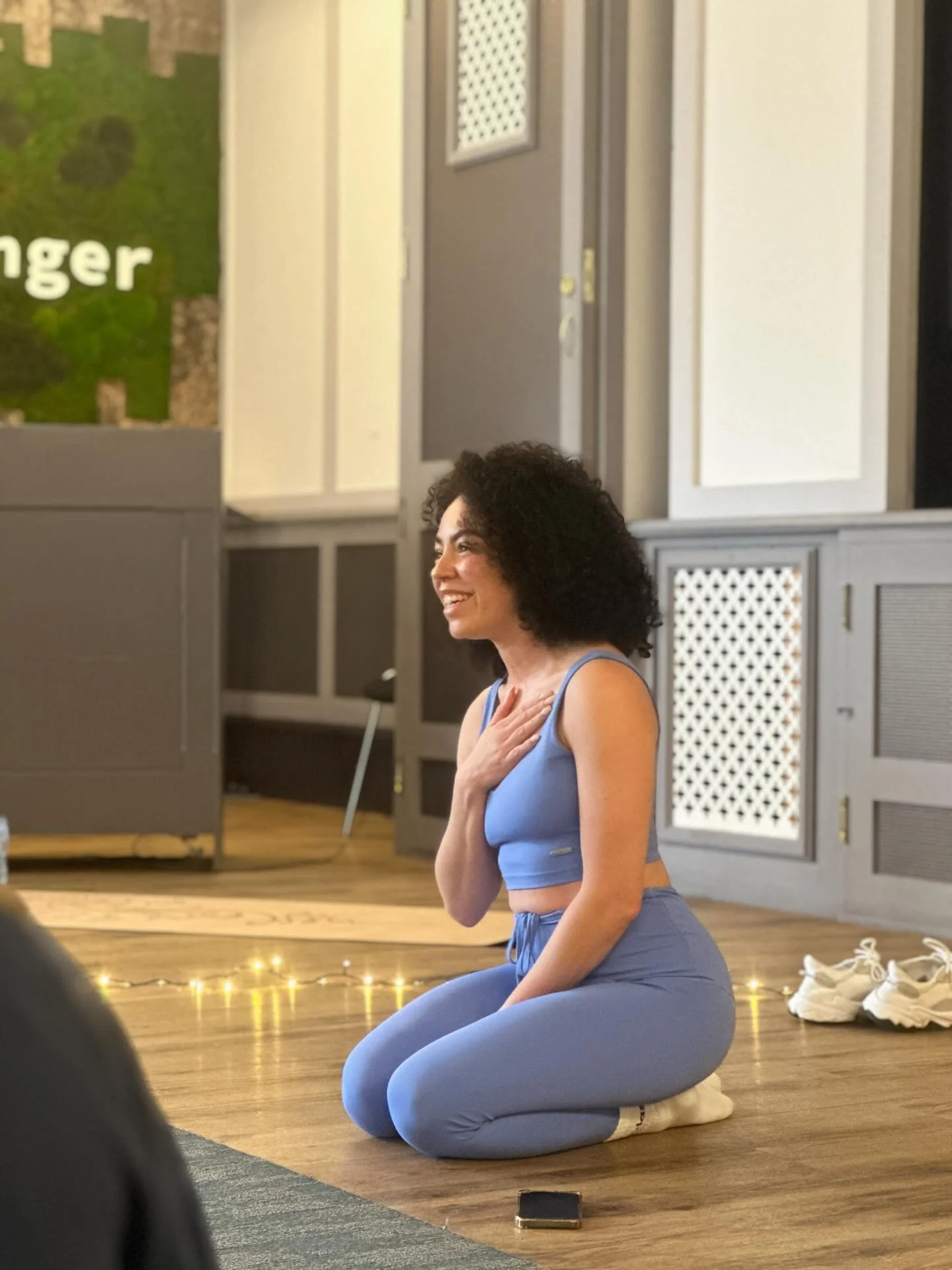 Woman with curly hair practicing yoga indoors, kneeling on a cushion, with her hand on her chest, smiling, in front of a gray cabinet and a green wall with white text.