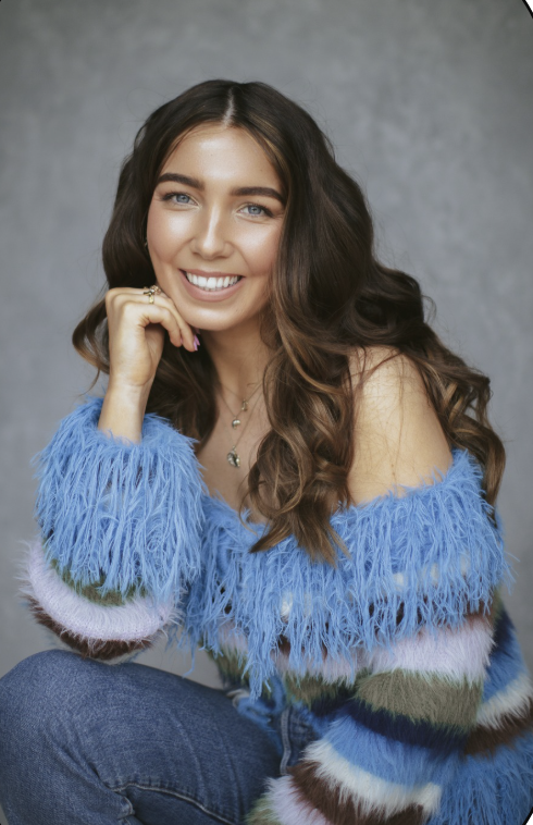 A young woman with long wavy brown hair, smiling, wearing a fuzzy multicolored off-the-shoulder sweater and jeans, sitting against a gray background.