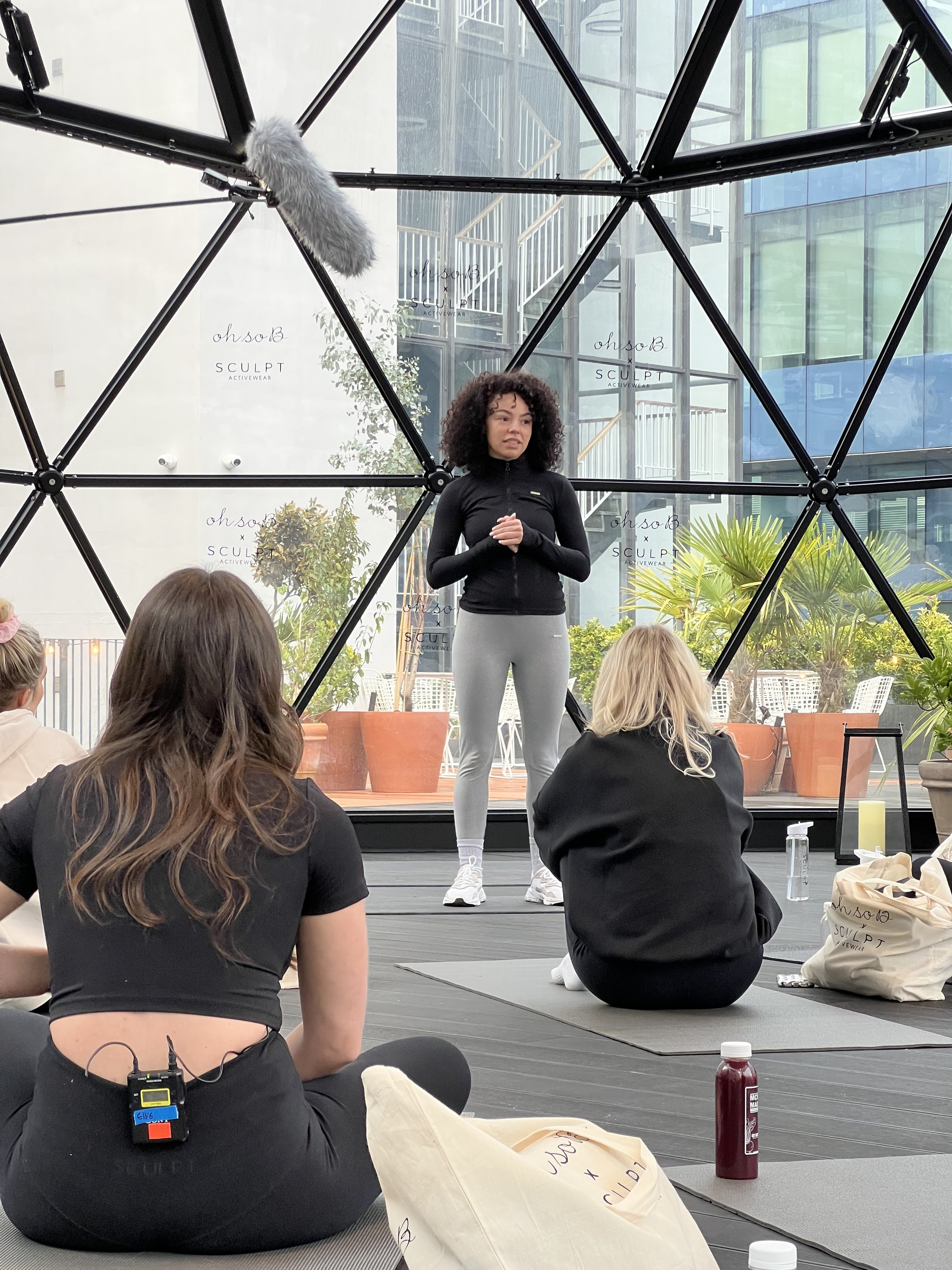 A woman in athletic clothing standing and speaking to a seated group of women in a fitness or wellness class inside a glass-walled building with plants and an urban exterior visible.