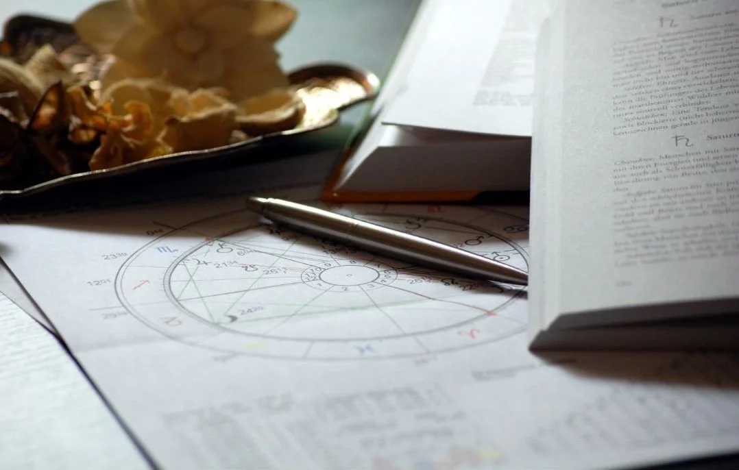 Open book, pen, astrology chart, scattered papers, and a dish with dried flowers or herbs on a desk.
